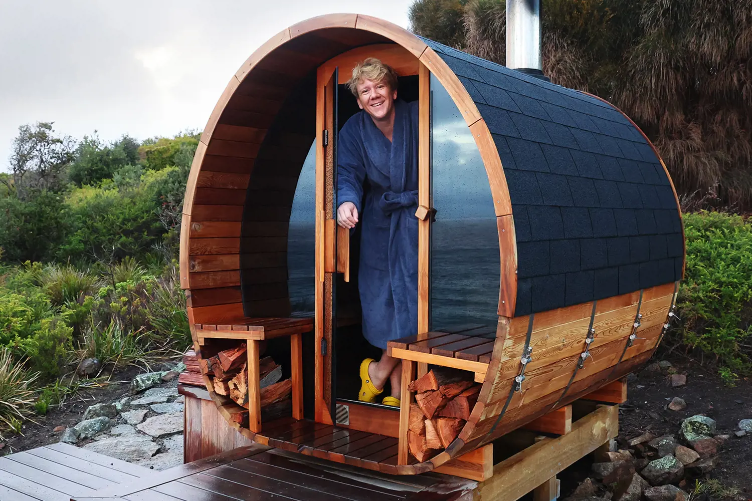 A man wearing a towel robe stands in the open doorway of a wooden drum-shaped sauna sitting a platform surrounded by bushlands behind and the sea in the background.