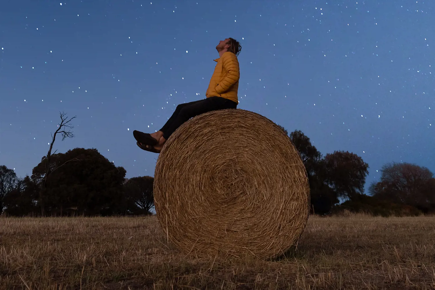 A man wearing a winter jacket sits on the top of a large cylindrical-shaped hay bale and looks up to the stars on a clear night.