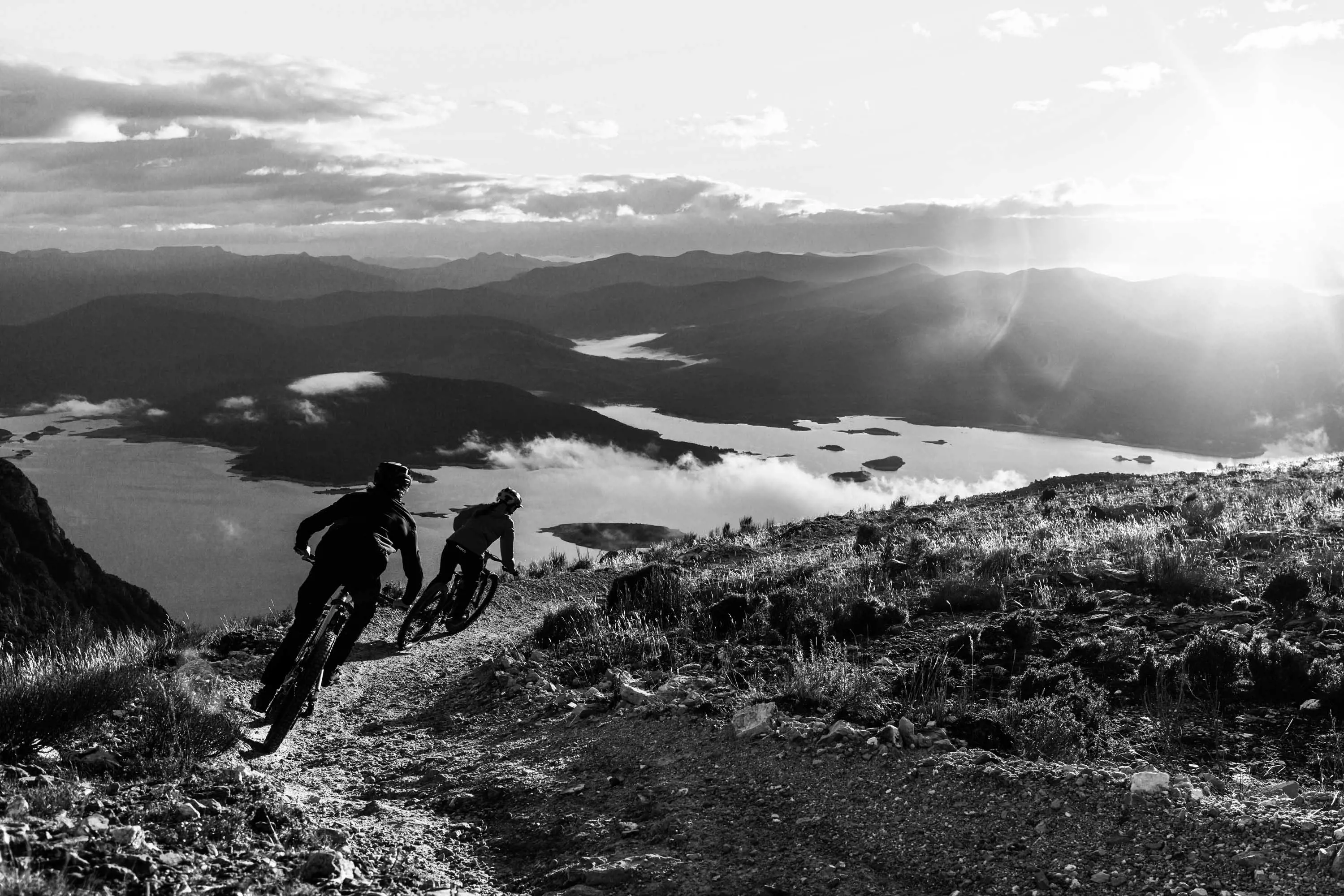 Two mountain bike riders riders lean into the corner of a gravel track looking down the side of a slope to a large lake in the distance.