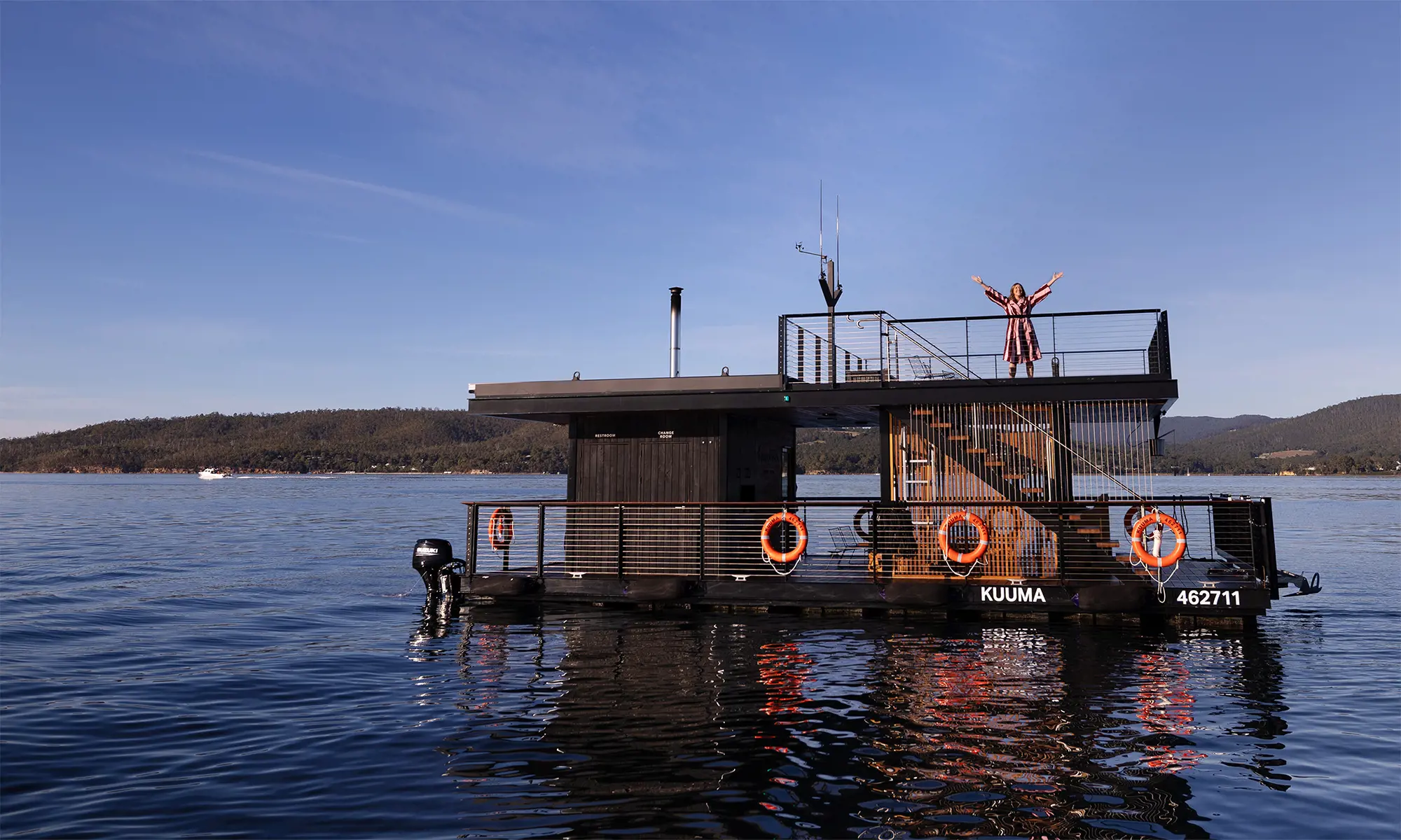 A woman standing on the rooftop deck of a modern black floating sauna with "Kuuma" written on the side, on a tranquil lake.