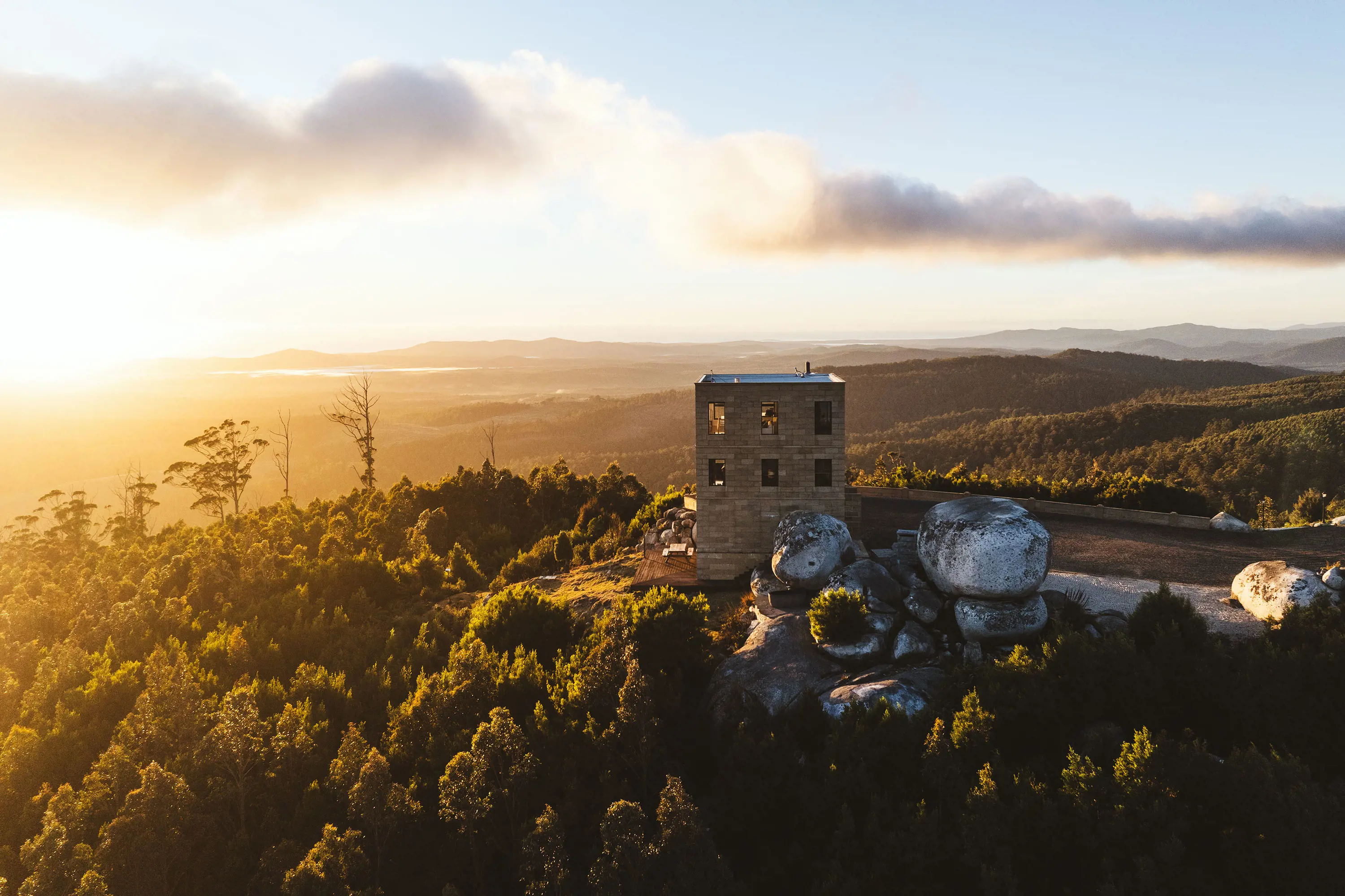 A square, solid 2-storey building with large windows all around sits atop a hill in a wild-looking landscape with huge boulders next to it.