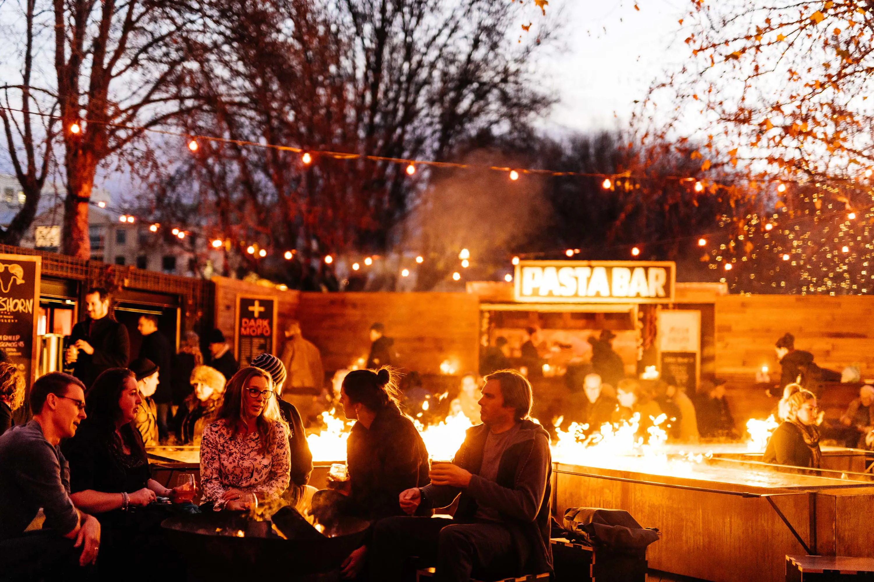 A small group of people sit near fire pits under trees in a port area of Hobart city early in the evening,