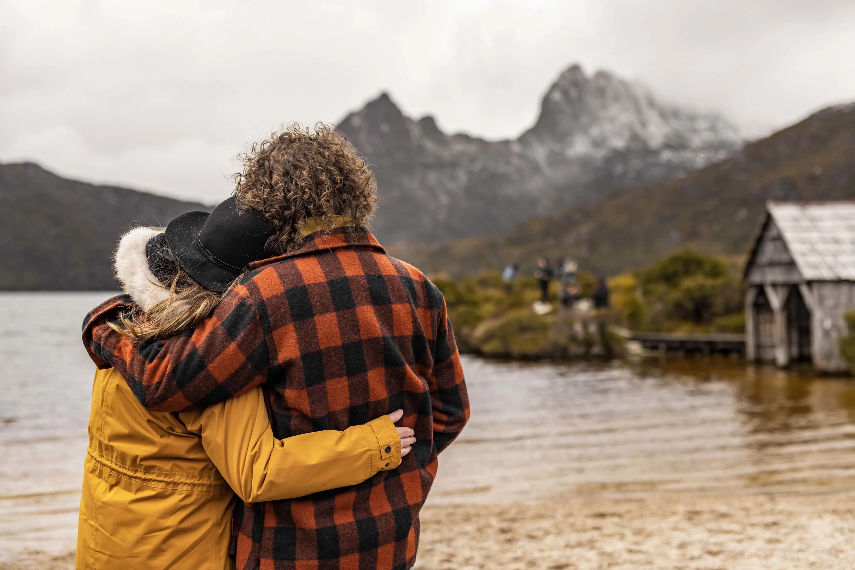 A couple stand at the edge of a clear water lake with a wooden boat shed and the large snow-capped peaks of a mountain in the background.