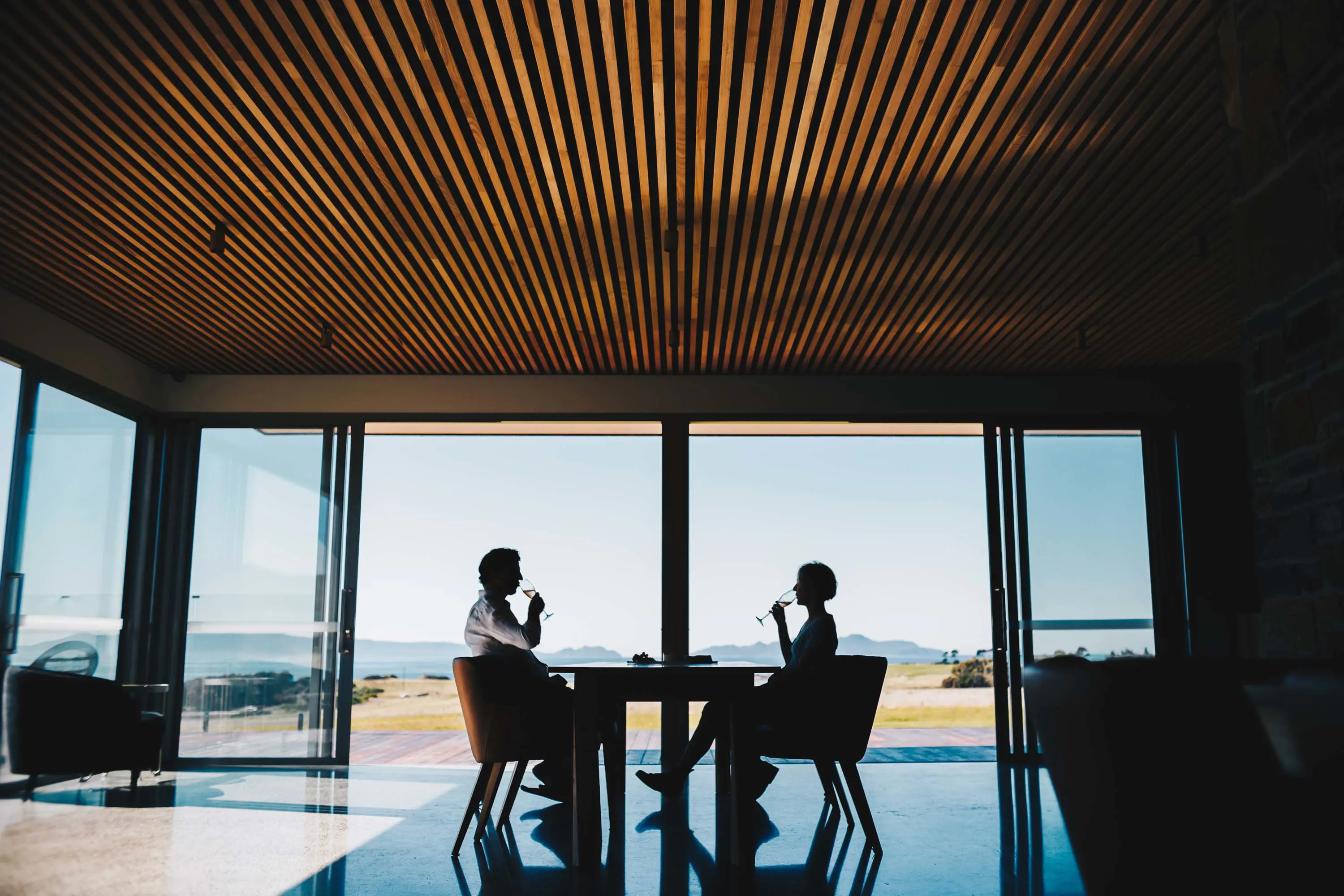 A couple drink wine at a table in a modern dining room with polished concrete floors and large floor-to-ceiling windows looking over low-lying mountains in the distance.