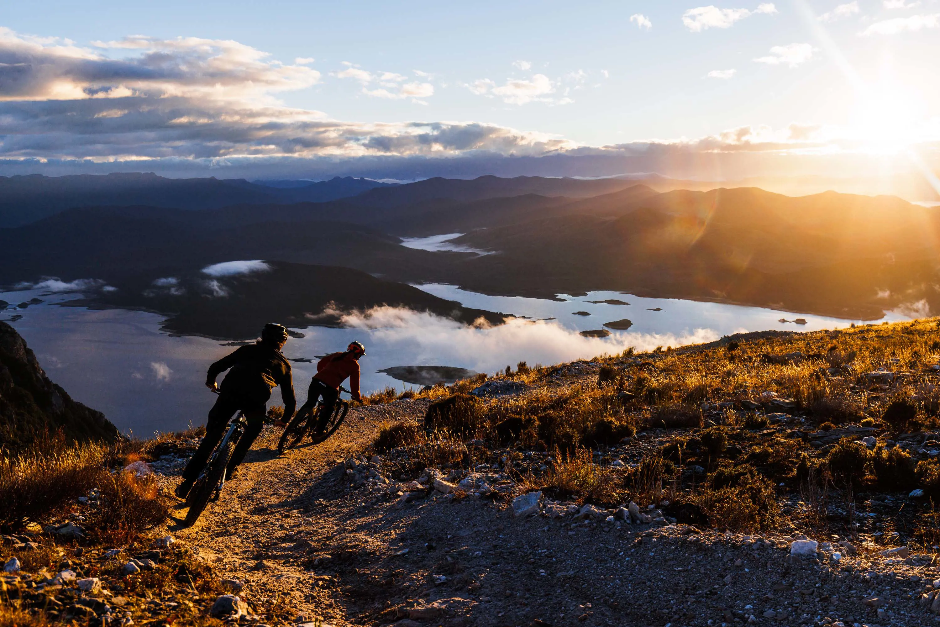 Two mountain bike riders turn a corner on a gravel track with sunset views of a valley shrouded in thick, white, low-lying cloud.