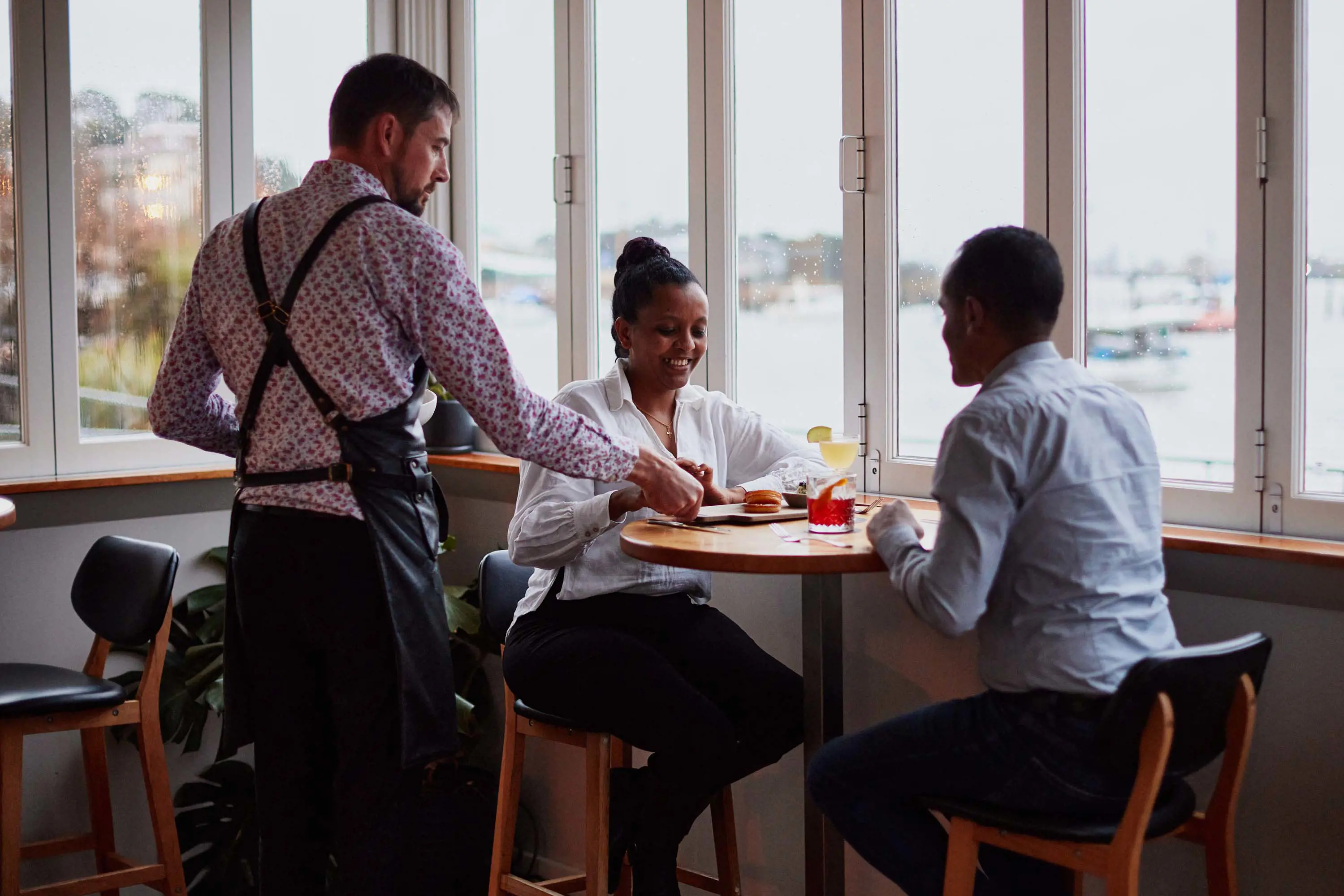 A waiter serves a couple sitting by windows looking out over water in a light and airy restaurant.