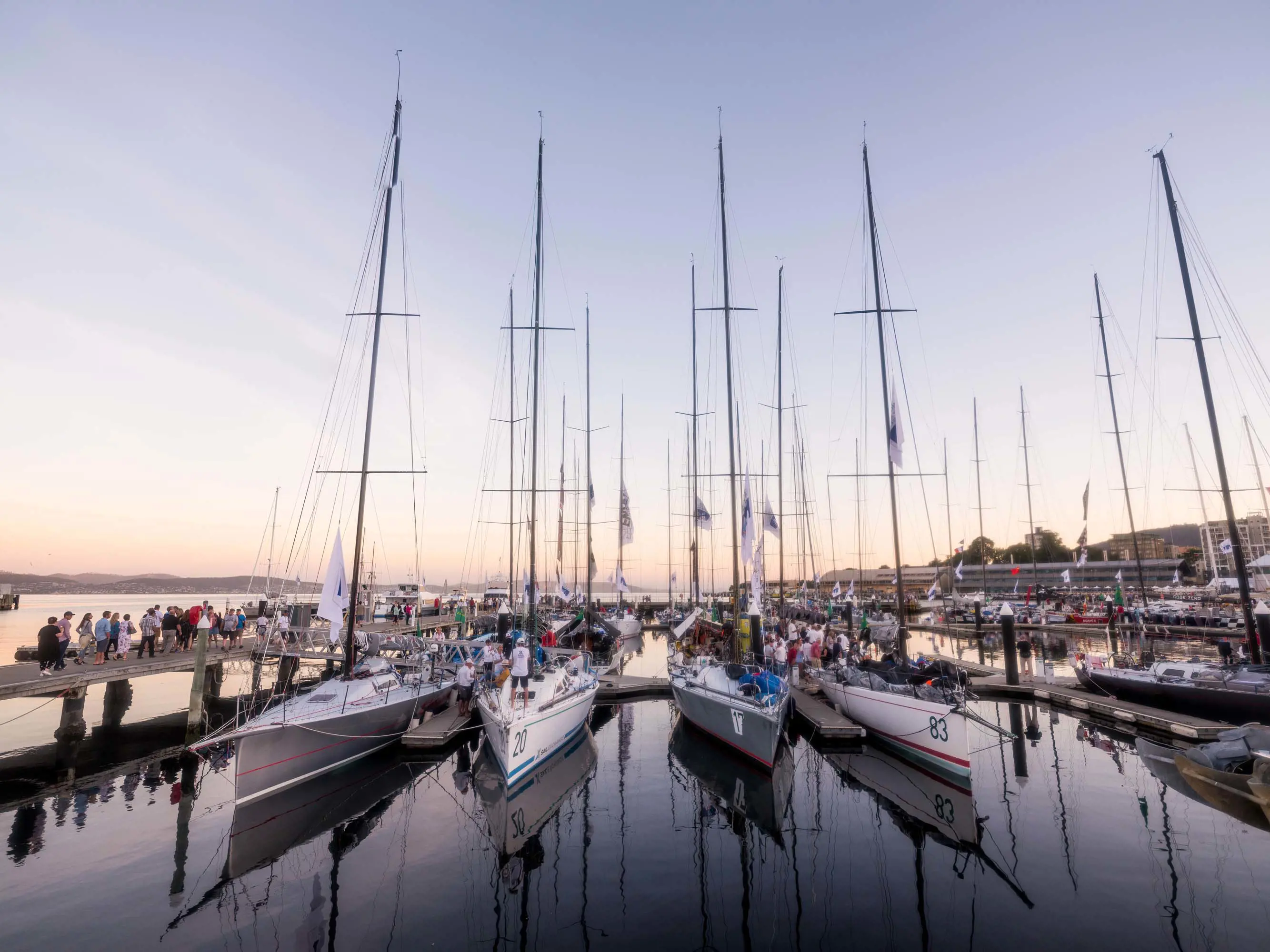 A row of racing yachts float in the port at the Hobart waterfront. Groups of people walk along piers to get a closer look.