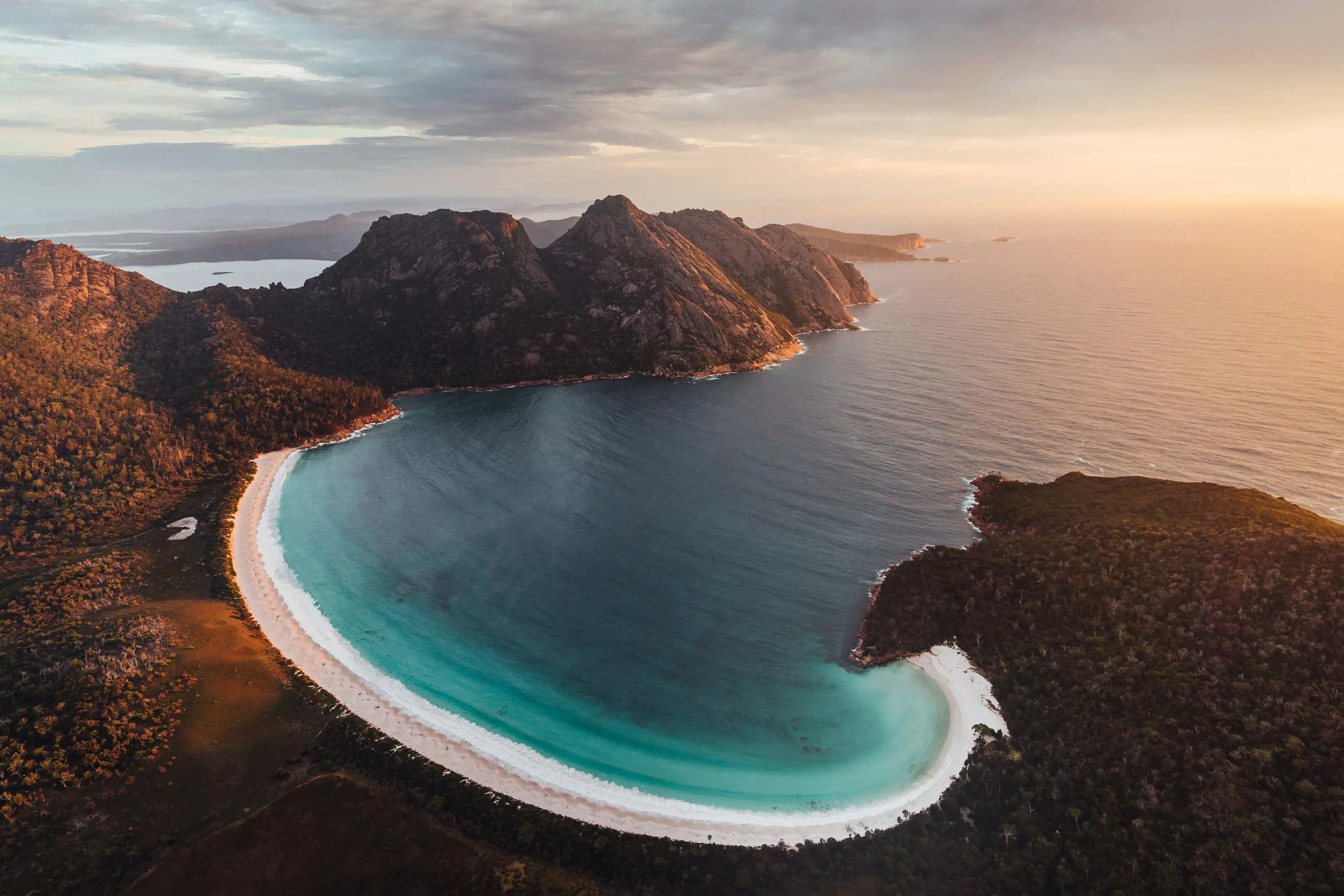 A secluded bay with a white sandy beach surrounded by forest and mountains at sunset.