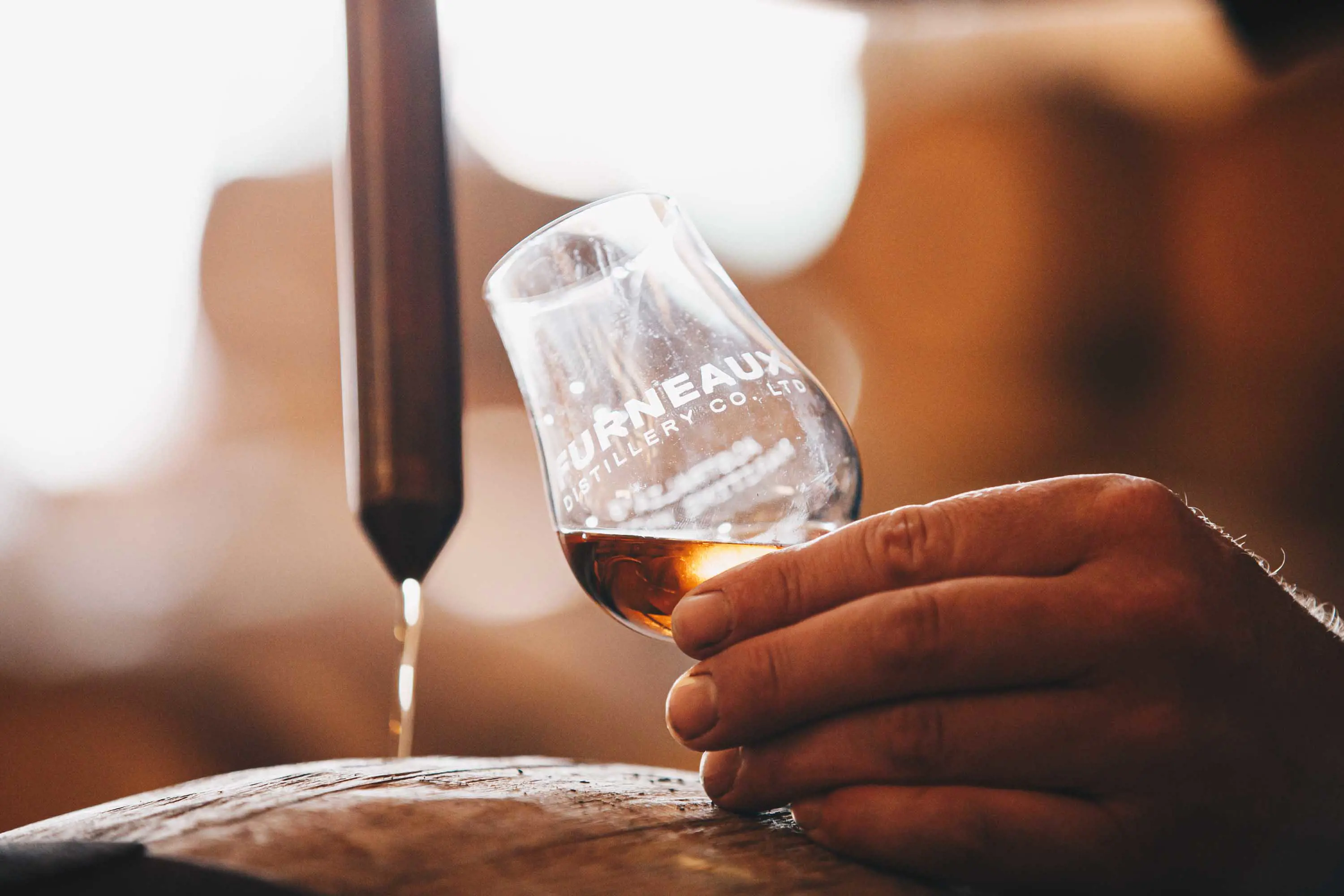 A man uses a long metal pipette and a small glass to check the flavour of whiskey taken from a wooden barrel.