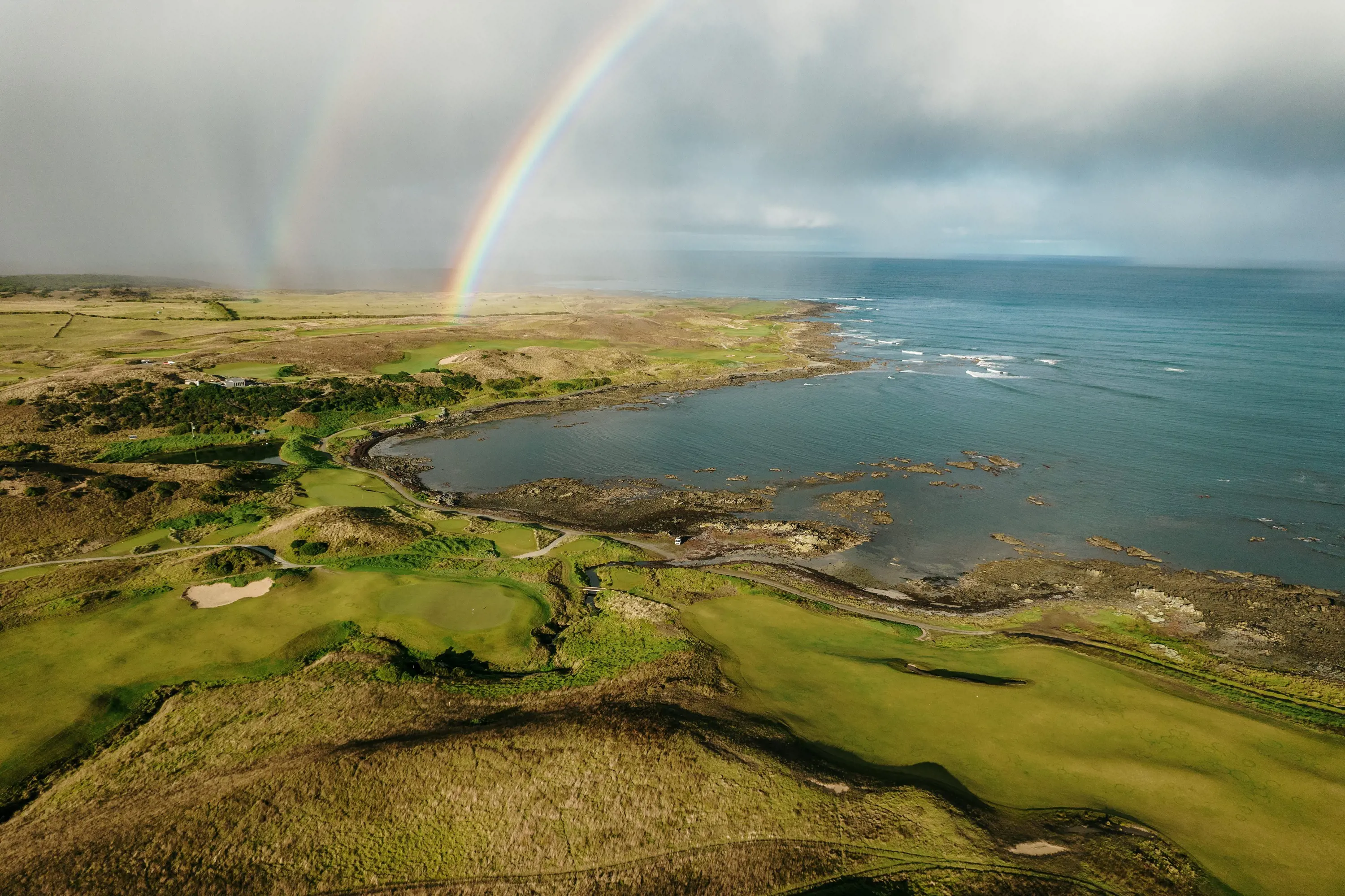 A golf course stretches along a coastline with wild seas and a rainbow stretching across the sky.