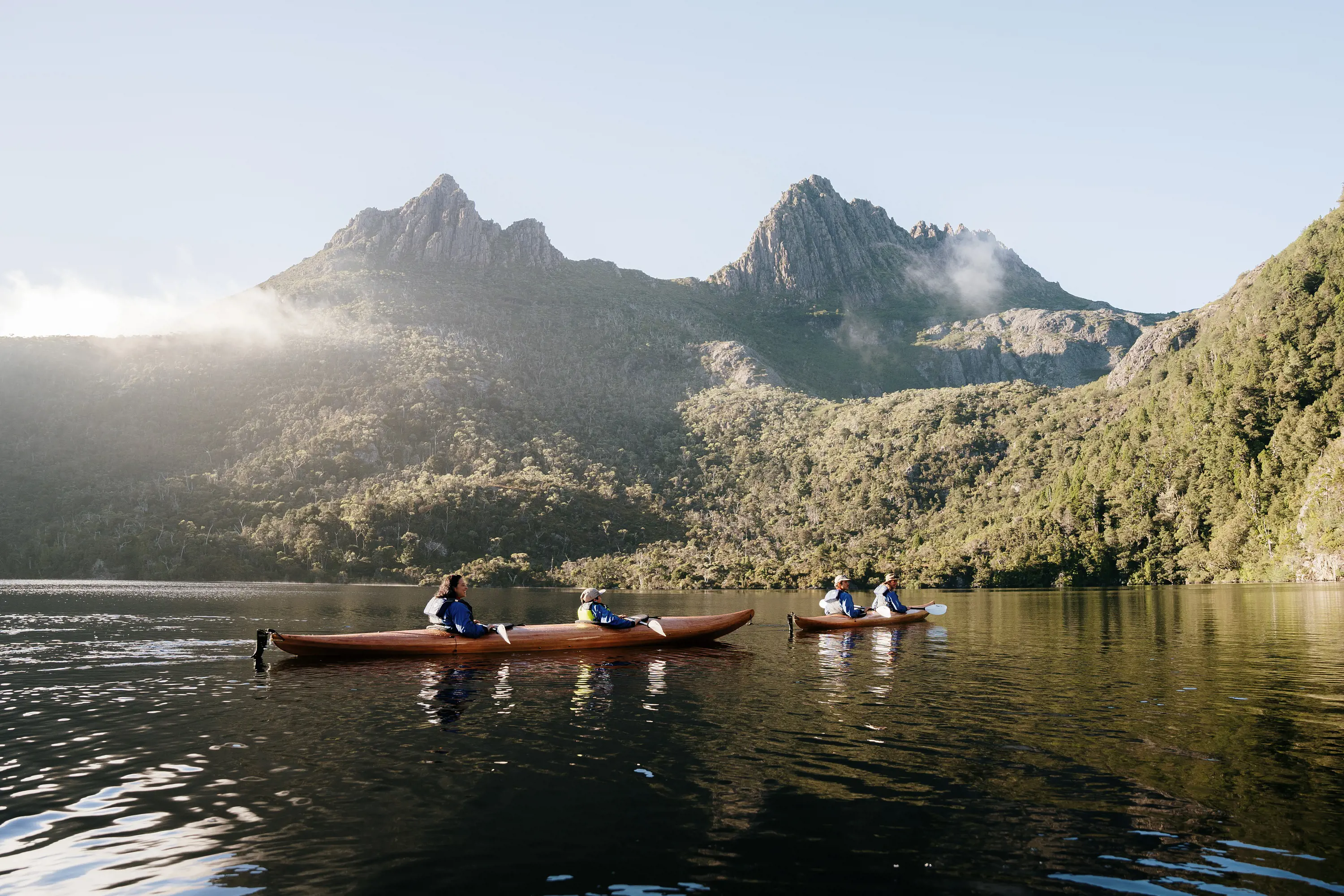 Two wooden canoes float on the calm waters of a lake with tall, rocky peaks of a mountain in the background on a clear day. 