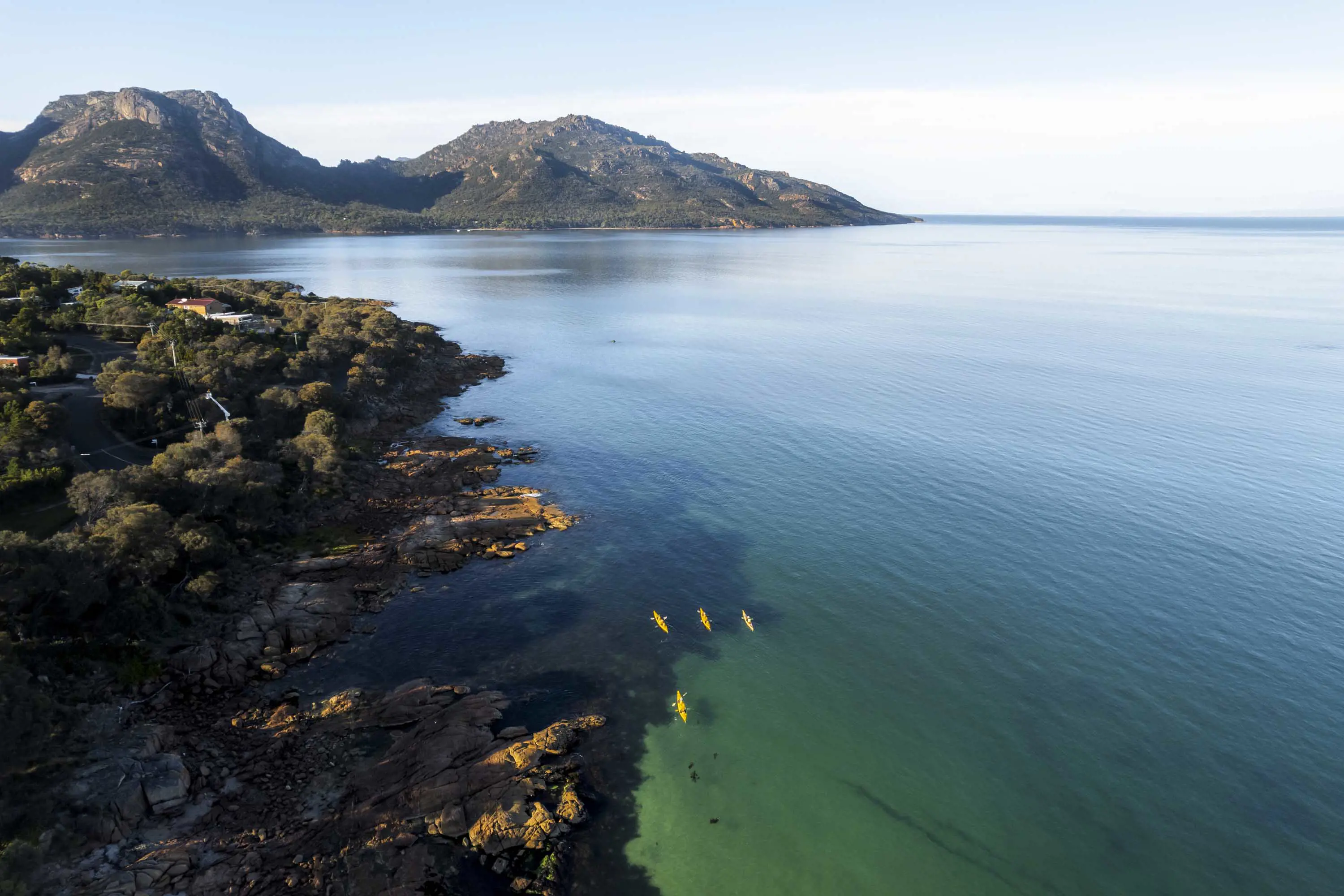 Yellow canoes round red rocks in light blue waters with mountains in the background on a clear sunny day.