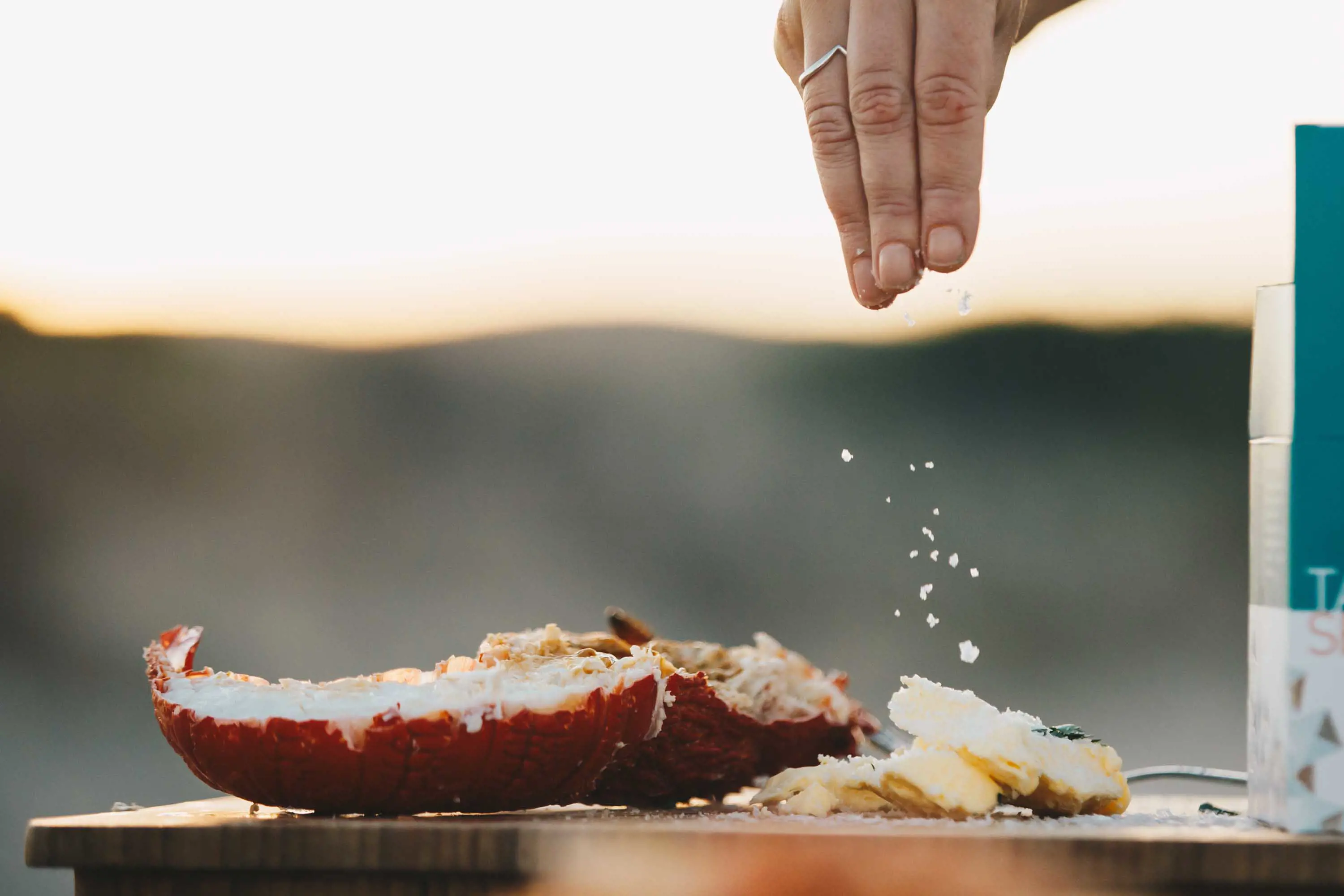 A hand sprinkles salt onto a cooked lobster tail sitting on a wood shopping block.