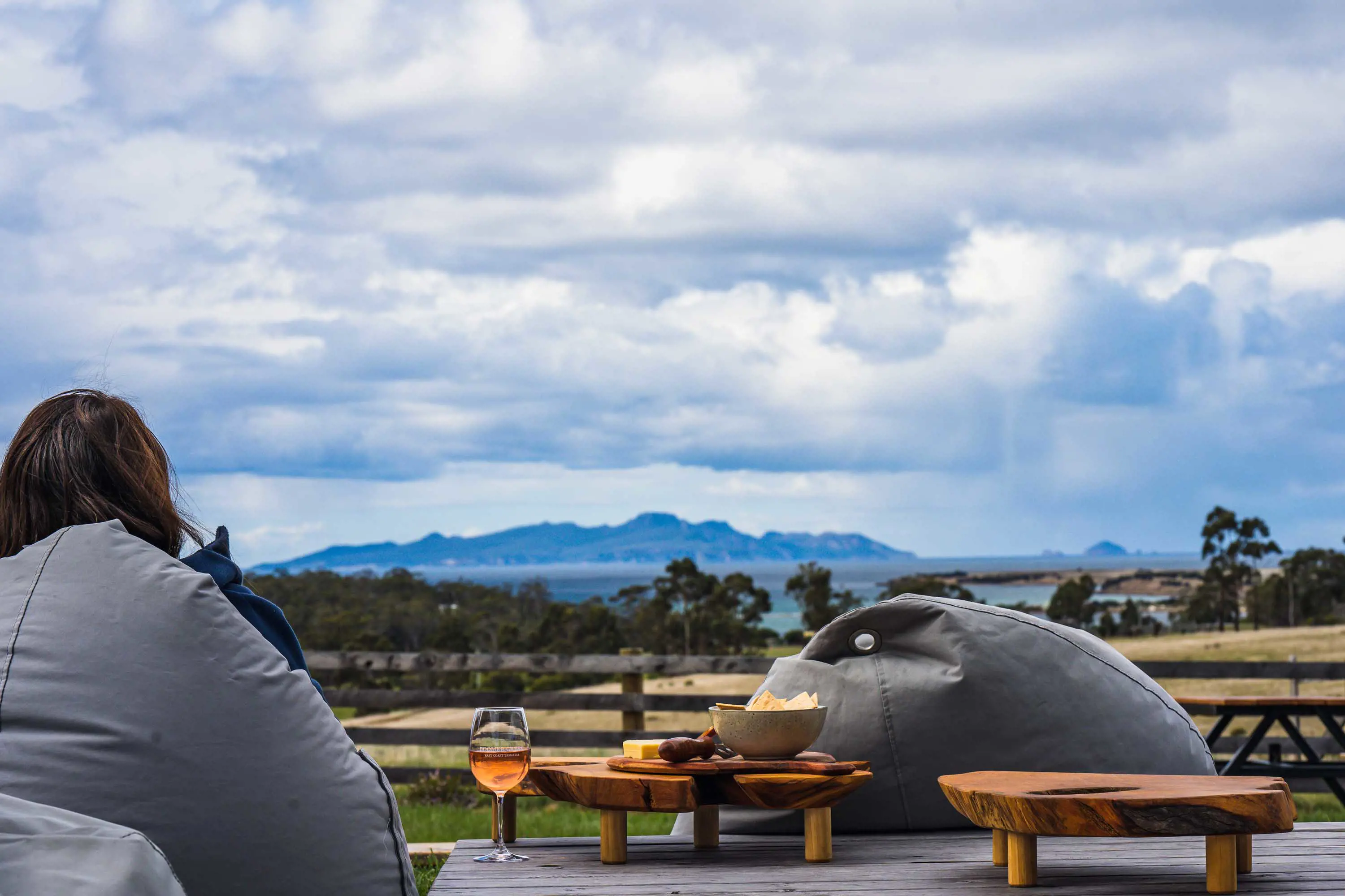 A person sits in a large, grey beanbag near small wooden tables with wine and food.