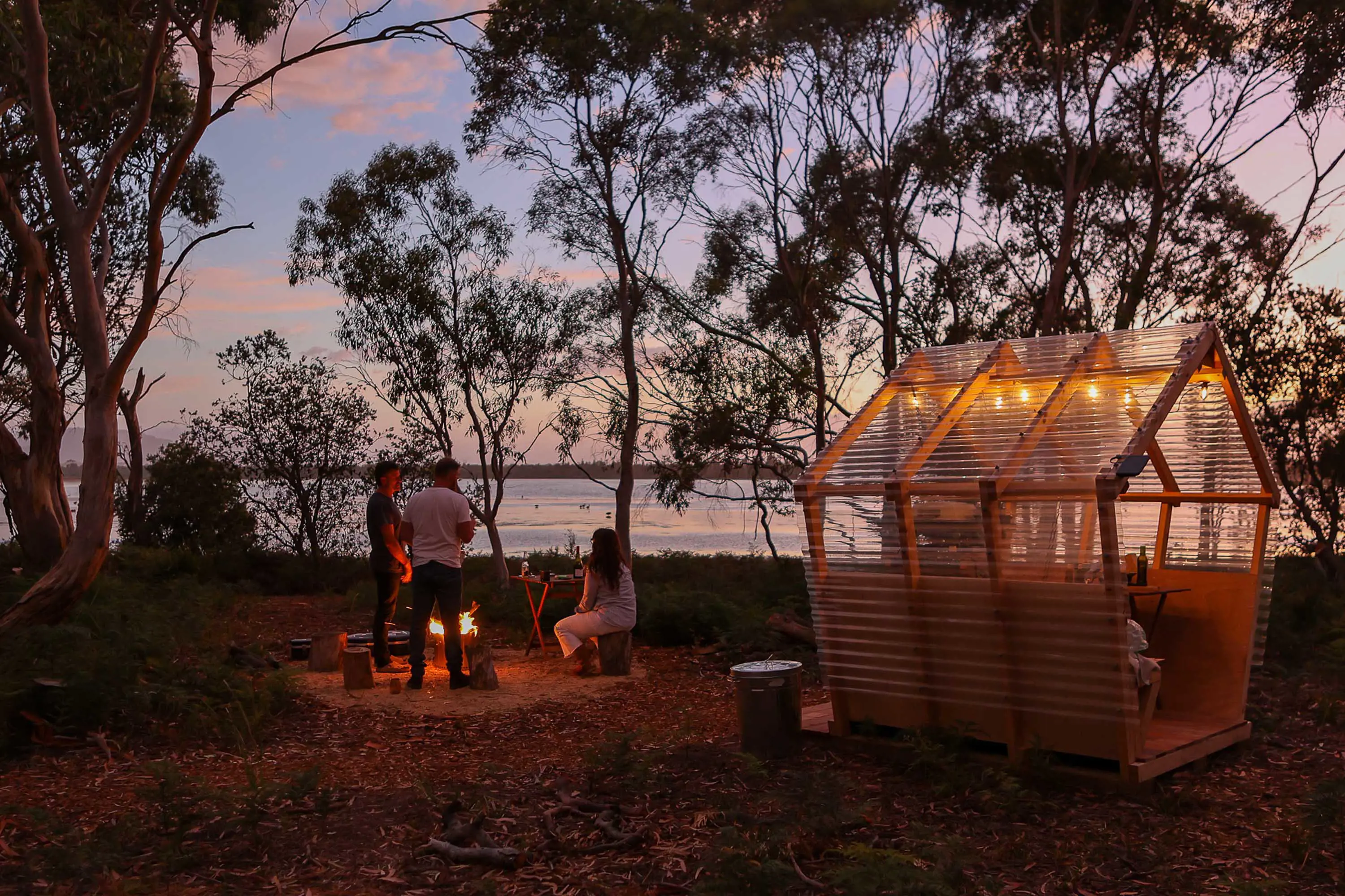 A group of people sit around a fire and talk with a small table with a clear roof shelter nearby.