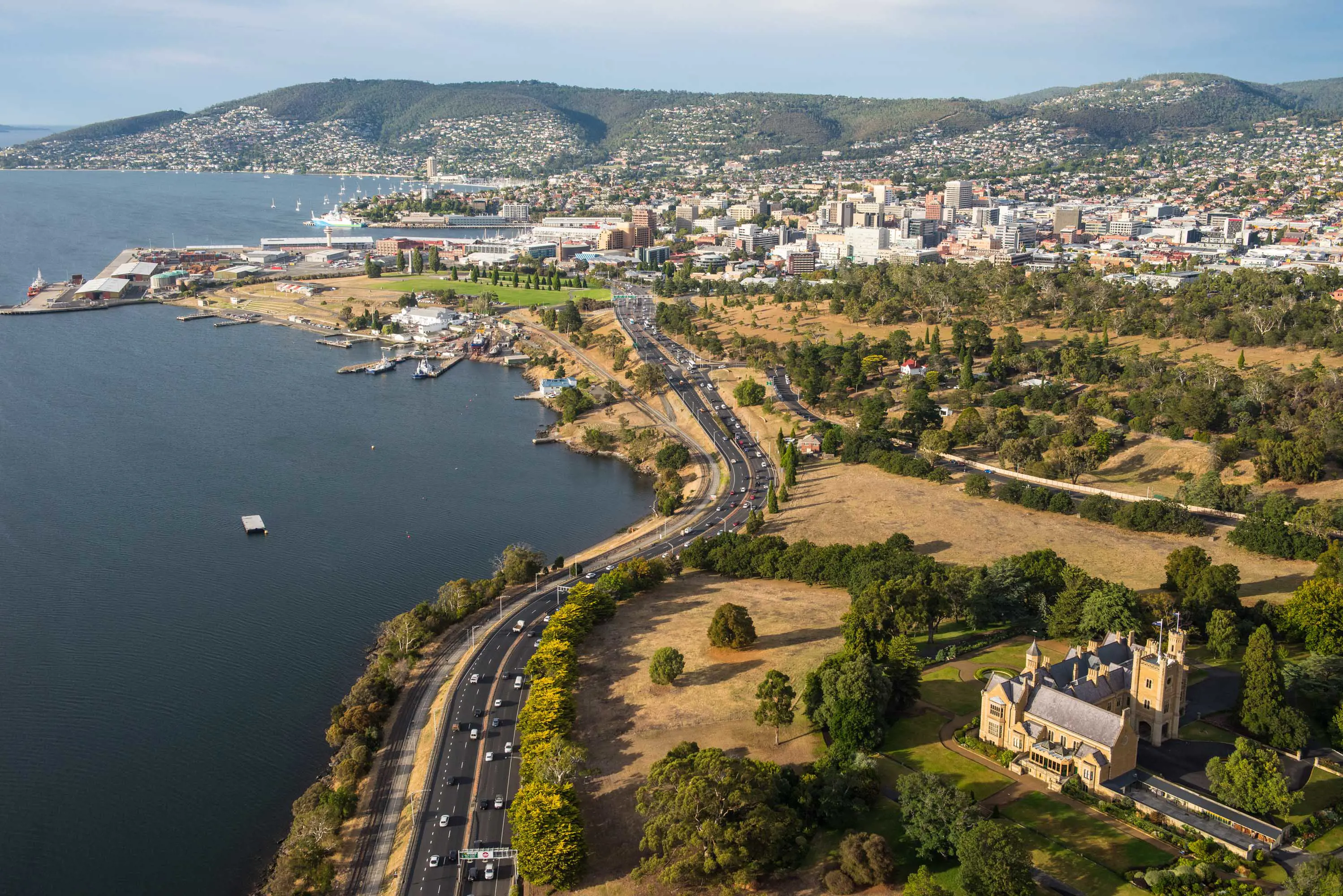 A 4-lane highway runs along the edge of a wide river at the edge of a city. A colonial era building stands on the top of a hill over looking the cars.