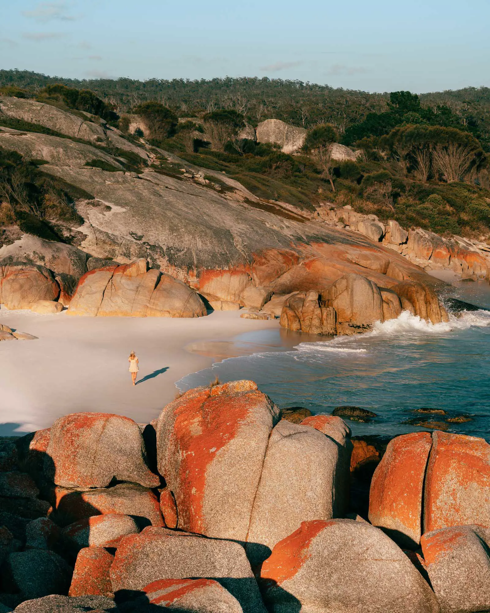 A woman wearing a broad-rimmed hat walks along the sand in a bay sheltered by red-lichen-covered rocks.