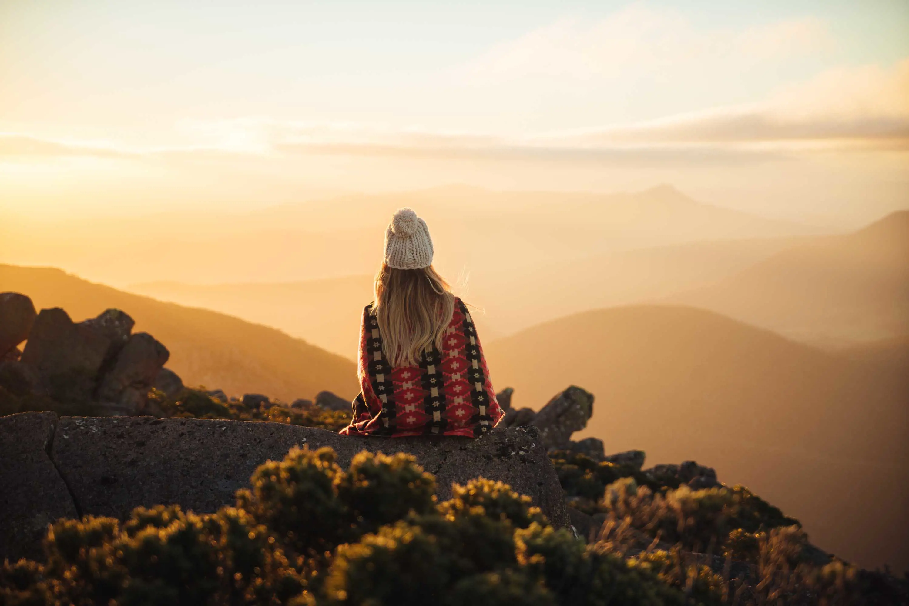 A woman wearing a beanie and a patterned blanket sits on a dolerite rock and looks over a valley in the warm sunlight of the afternoon.