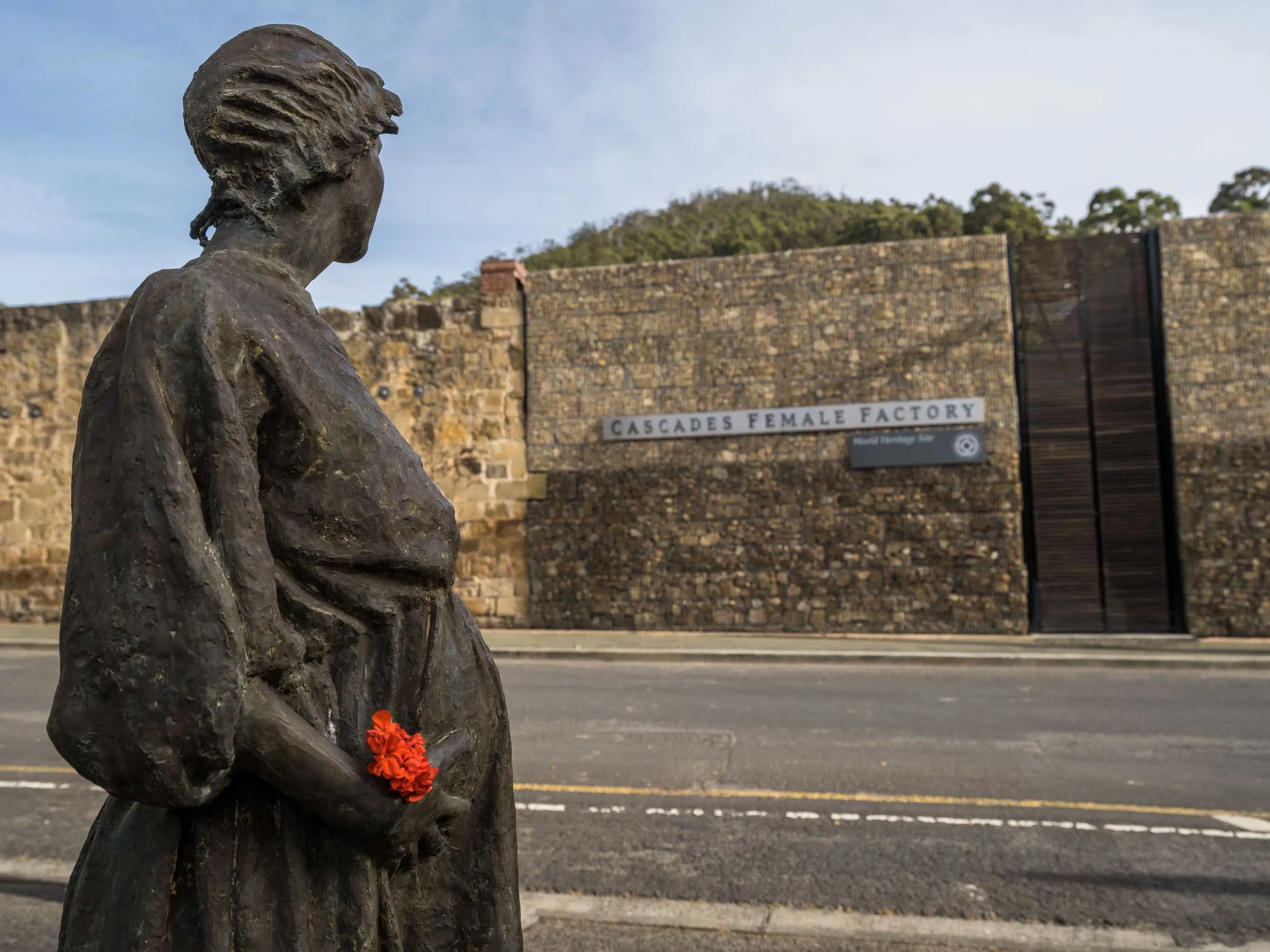 A bronze statue of a woman in early 19th century clothing stands on the side of a road opposite the large stone wall entrance to a historic site.