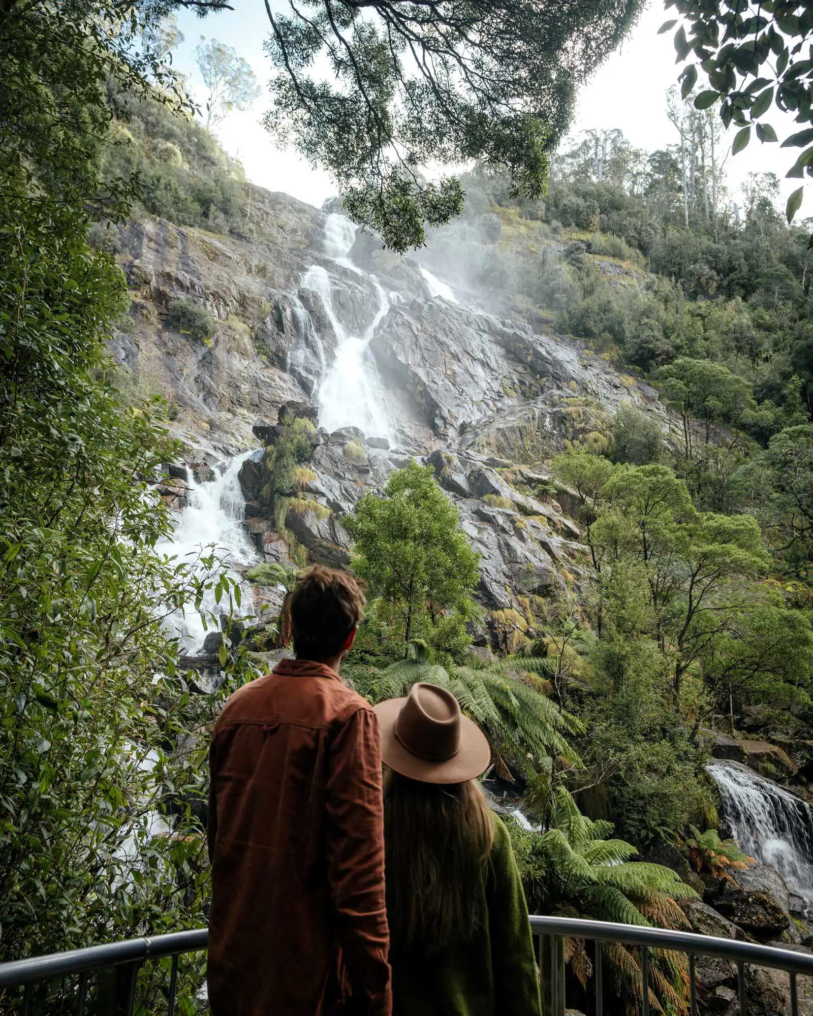 A couple stand at the base of a tall waterfall, that spills over the rocky outcrops of a cliff face above them.