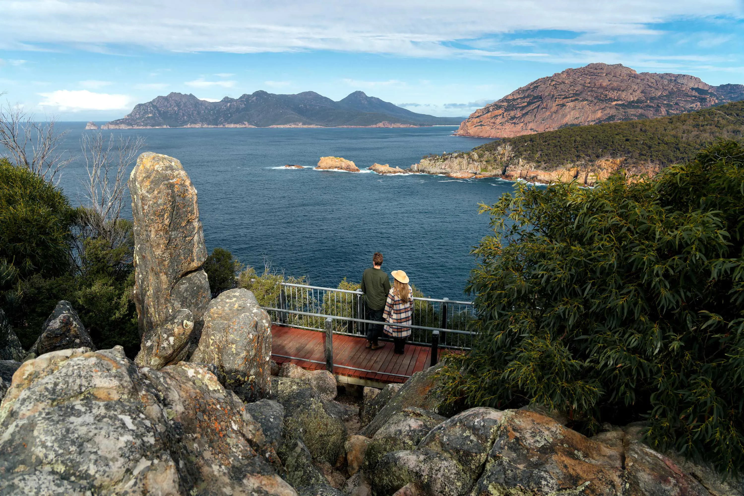 A young couple stand on the boardwalks on the edge of a coastal cliff looking out over water to a point on the horizon.