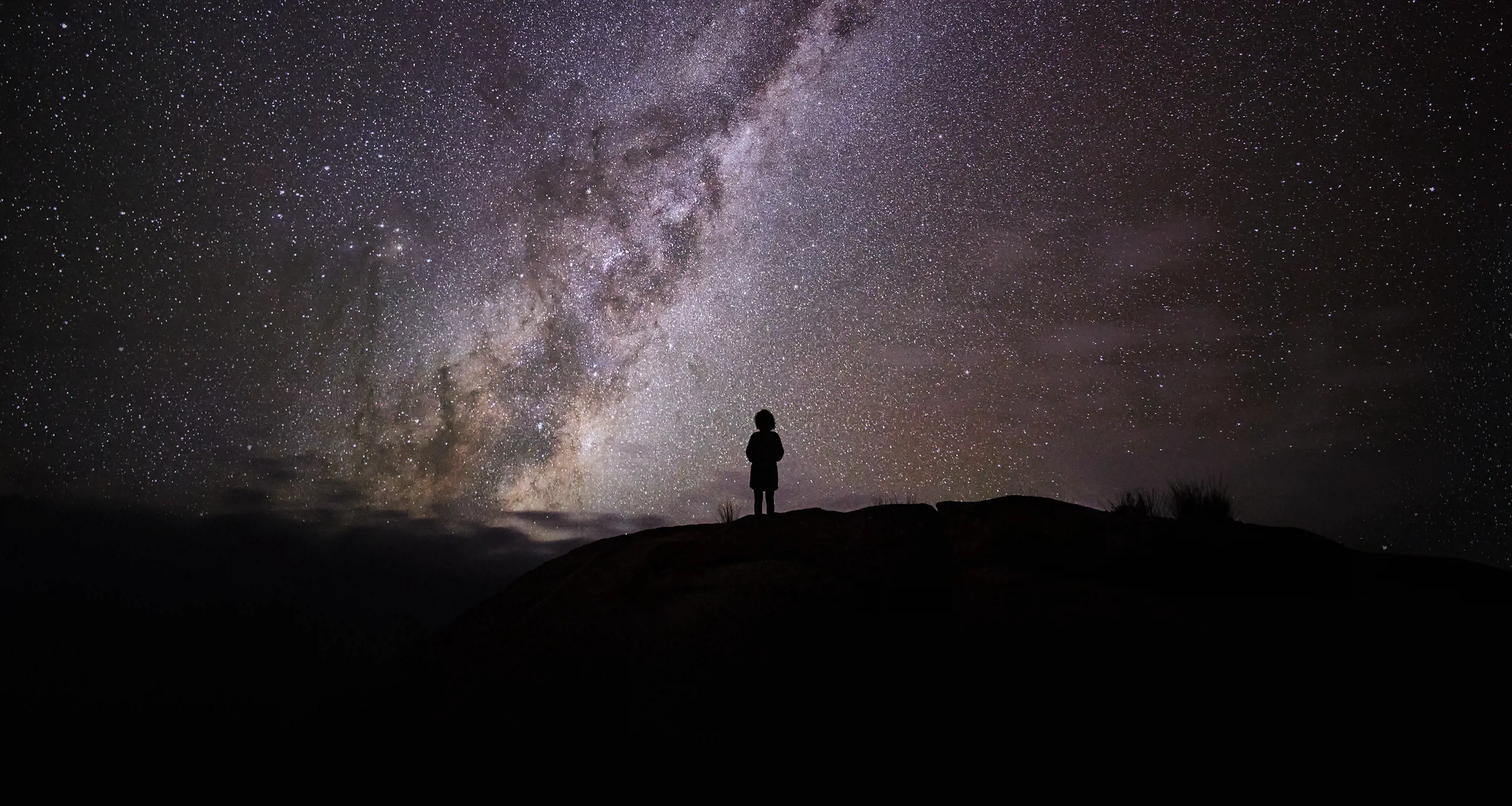 The milky way is clearly seen in hues of purple and white in the night sky. A silhouette of a person stands in the foreground.