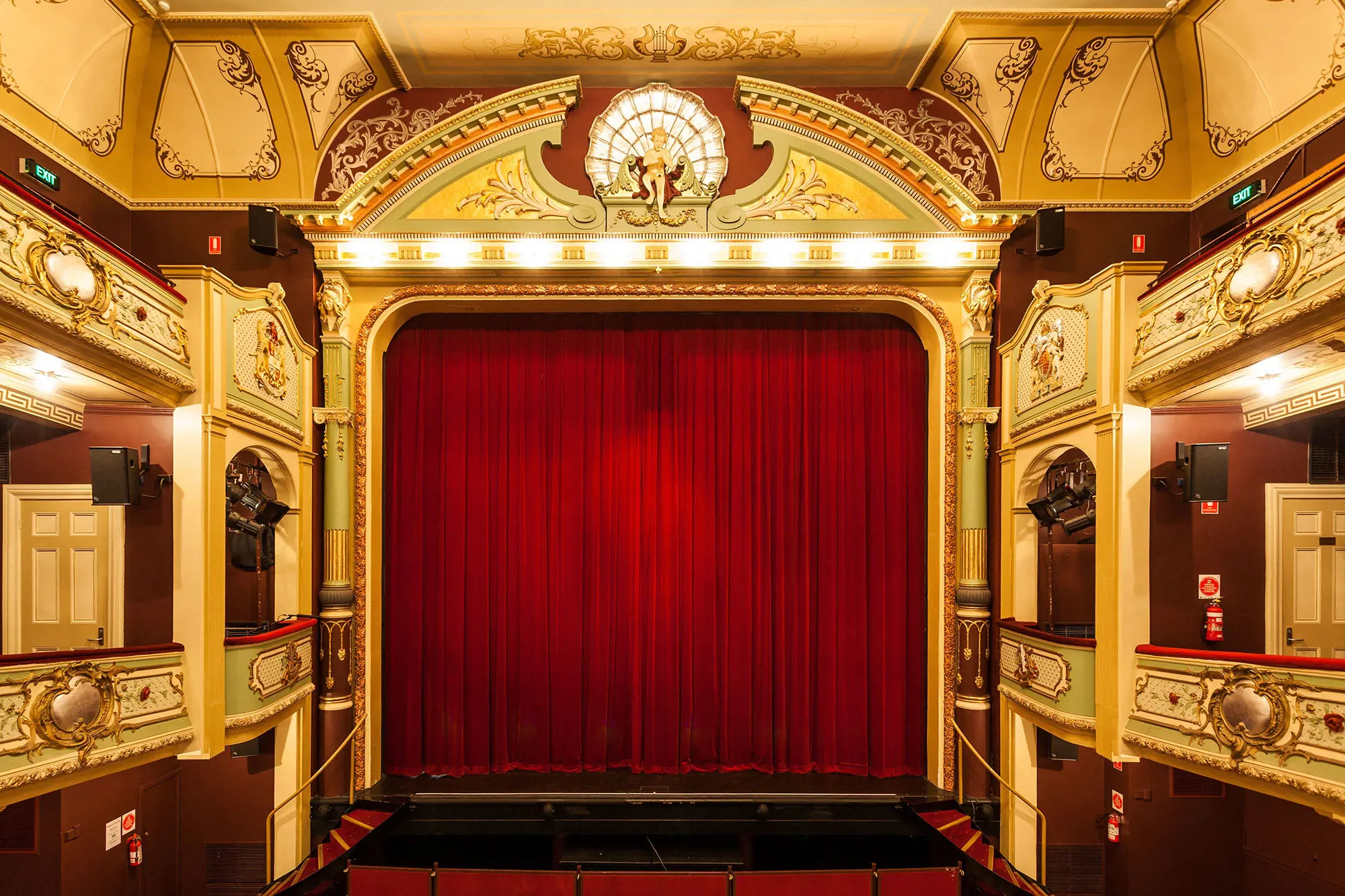 The interior of a richly-decorated 1830s-style theatre. There are several levels wrapping around the audience space and a thick red curtain is down over the stage.