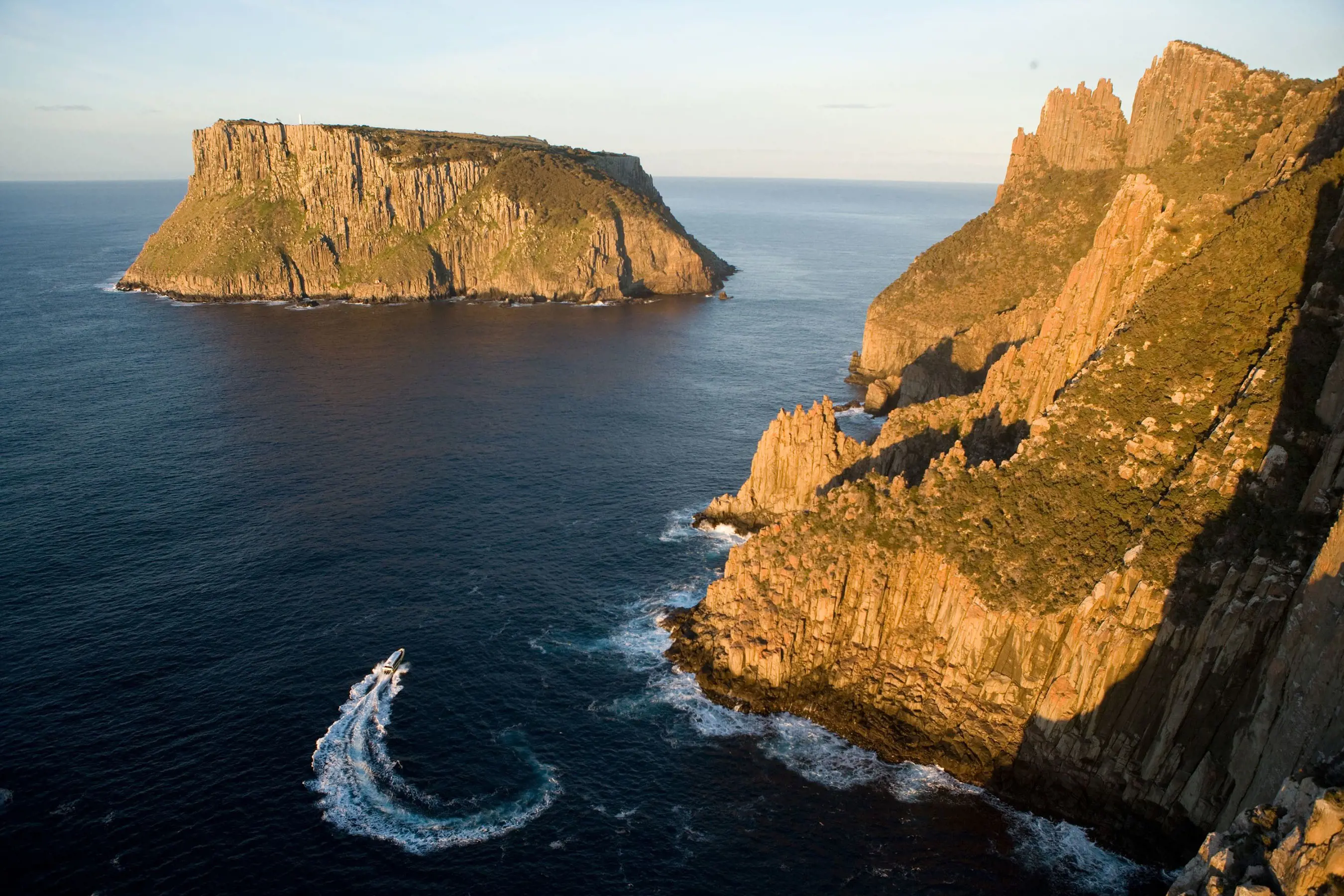 An aerial view of steep cliffs rising out of the water, and a small island with cliffs equally steep. In the ocean below, a sleek boat jets through the water.