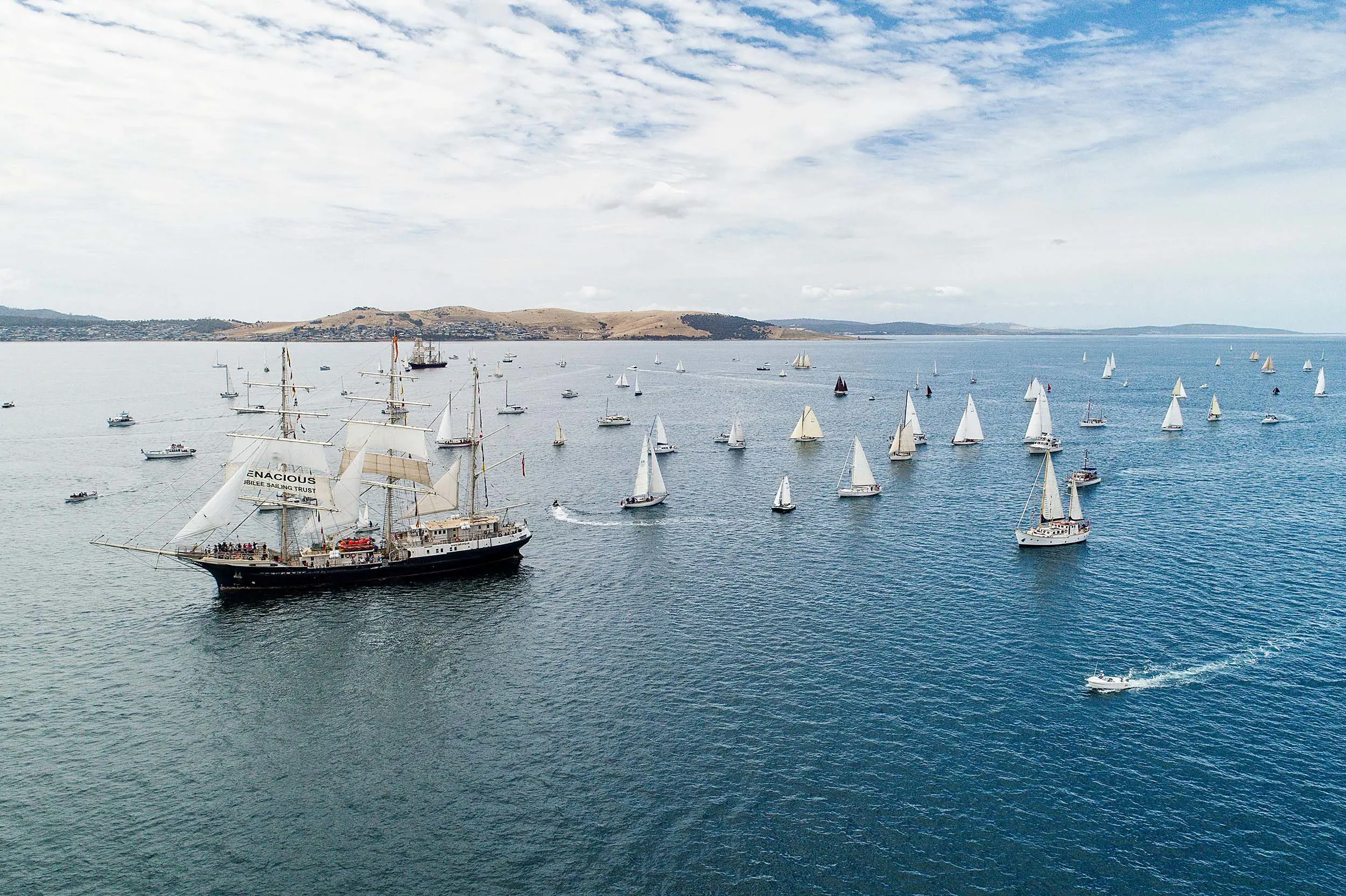 The calm waters of a river are dotted with boats, particularly a large, three-masted wooden sailing ship, and many small yachts.