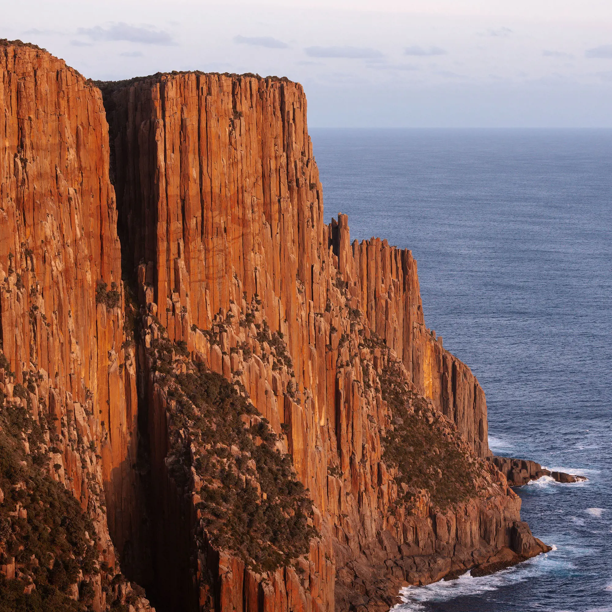 Enormous steep cliffs descend down into the dark ocean waters below. The cliffs seem almost orange in the sunset's glow.