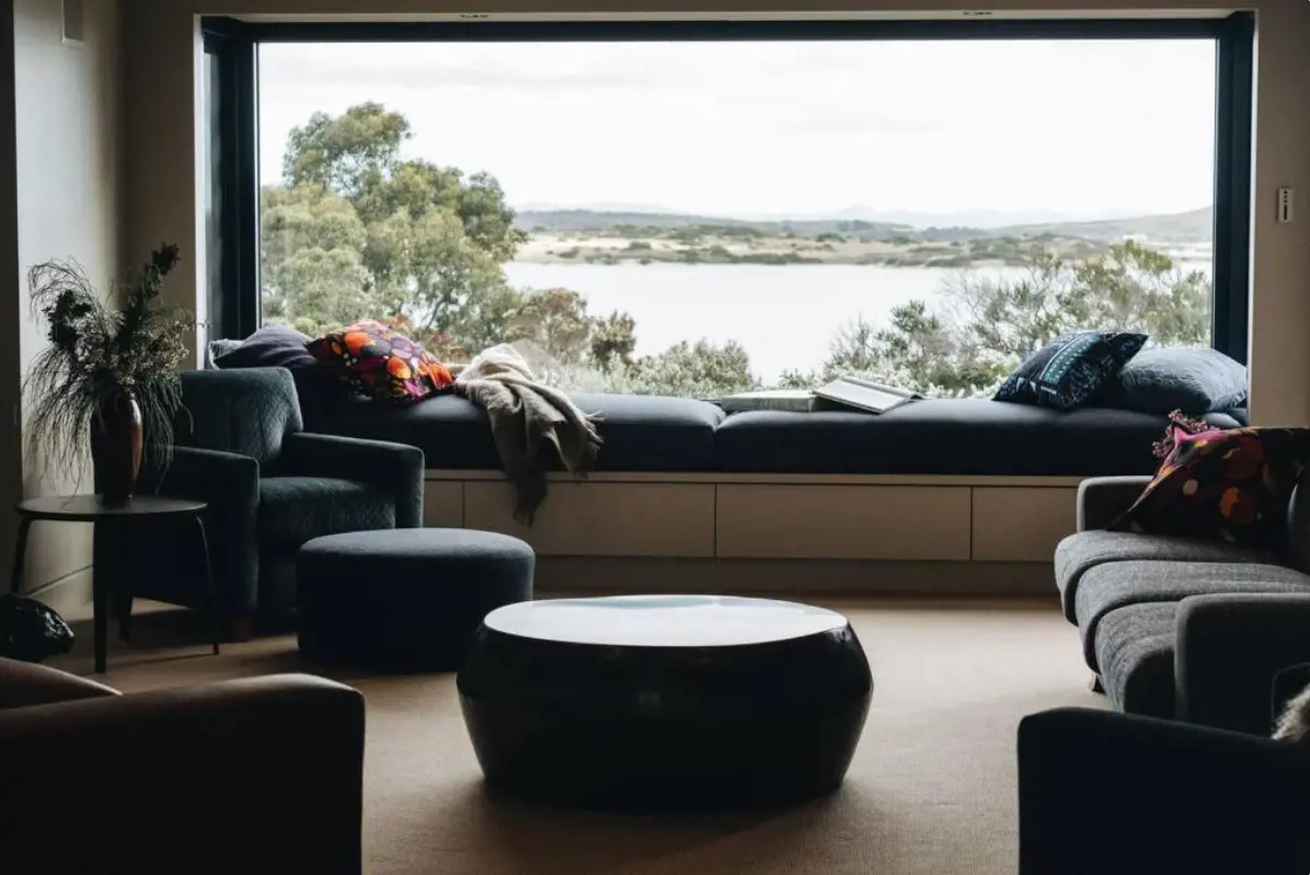 A window runs the length of a stylish living room, above a built-in window couch. Outside are views of the bush and a river. 