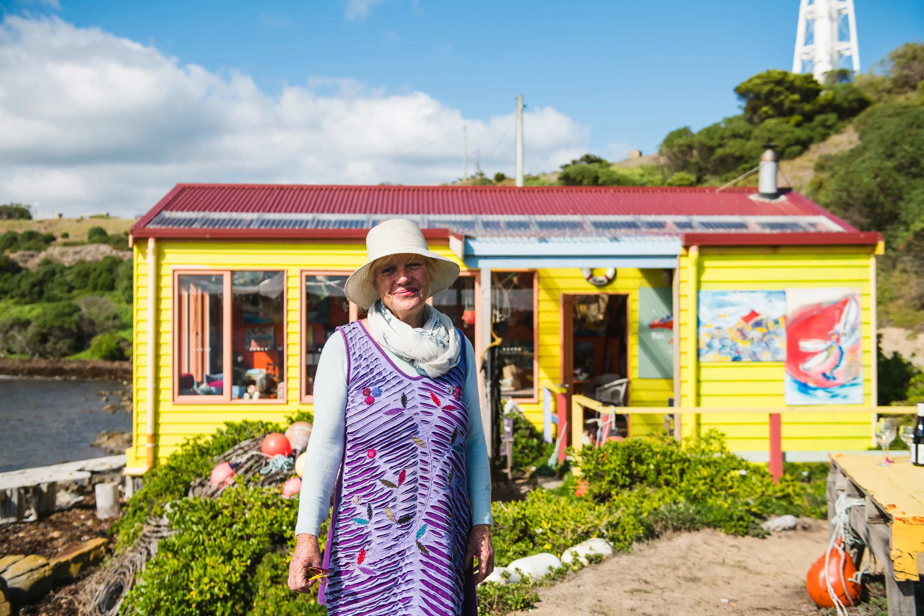 Caroline Kinonmonth stands in front of the Restaurant With No Food, a yellow, weather board boatshed looking out over water.