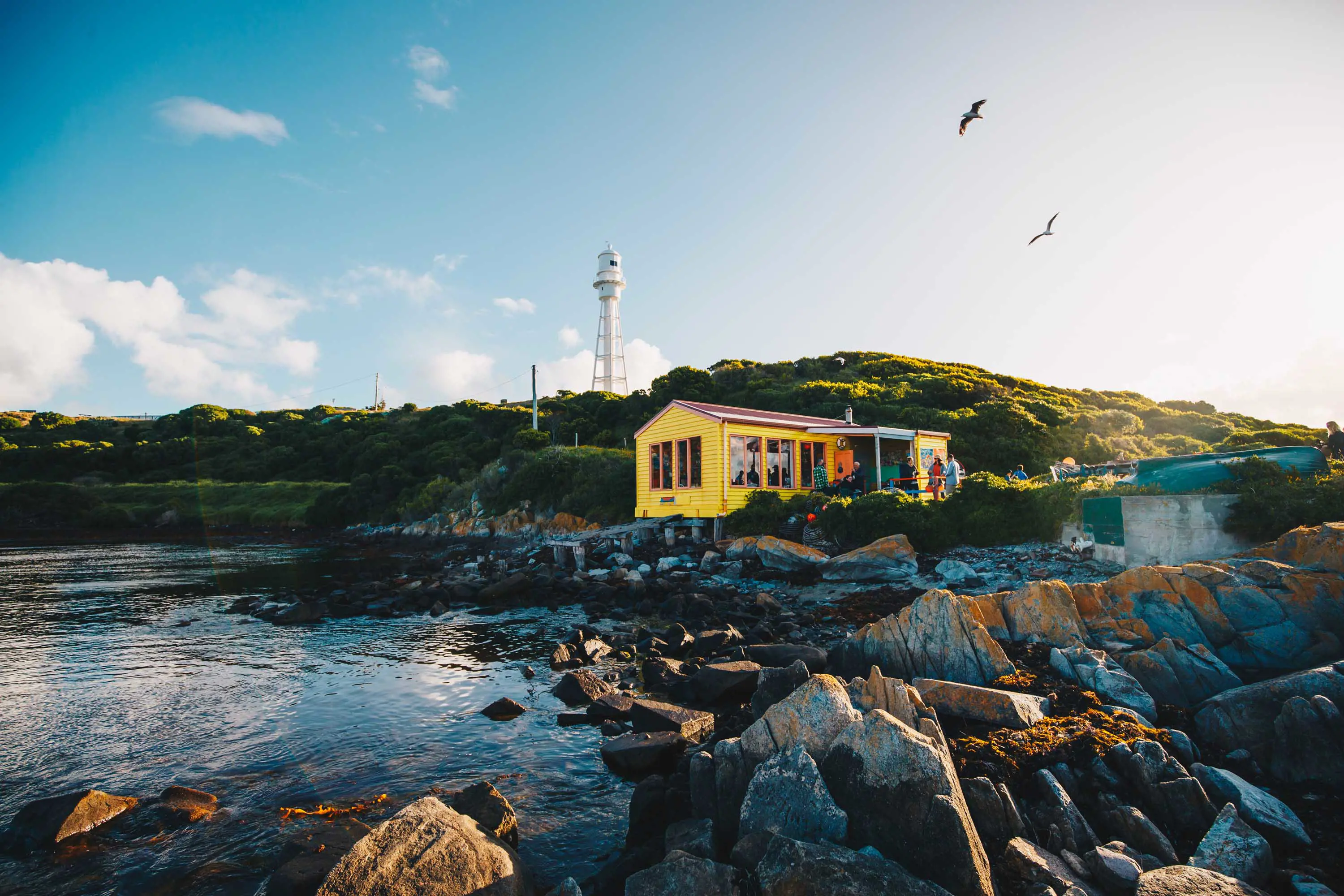 A yellow weatherboard house stands on a stony pier looking out over the water of a bay.