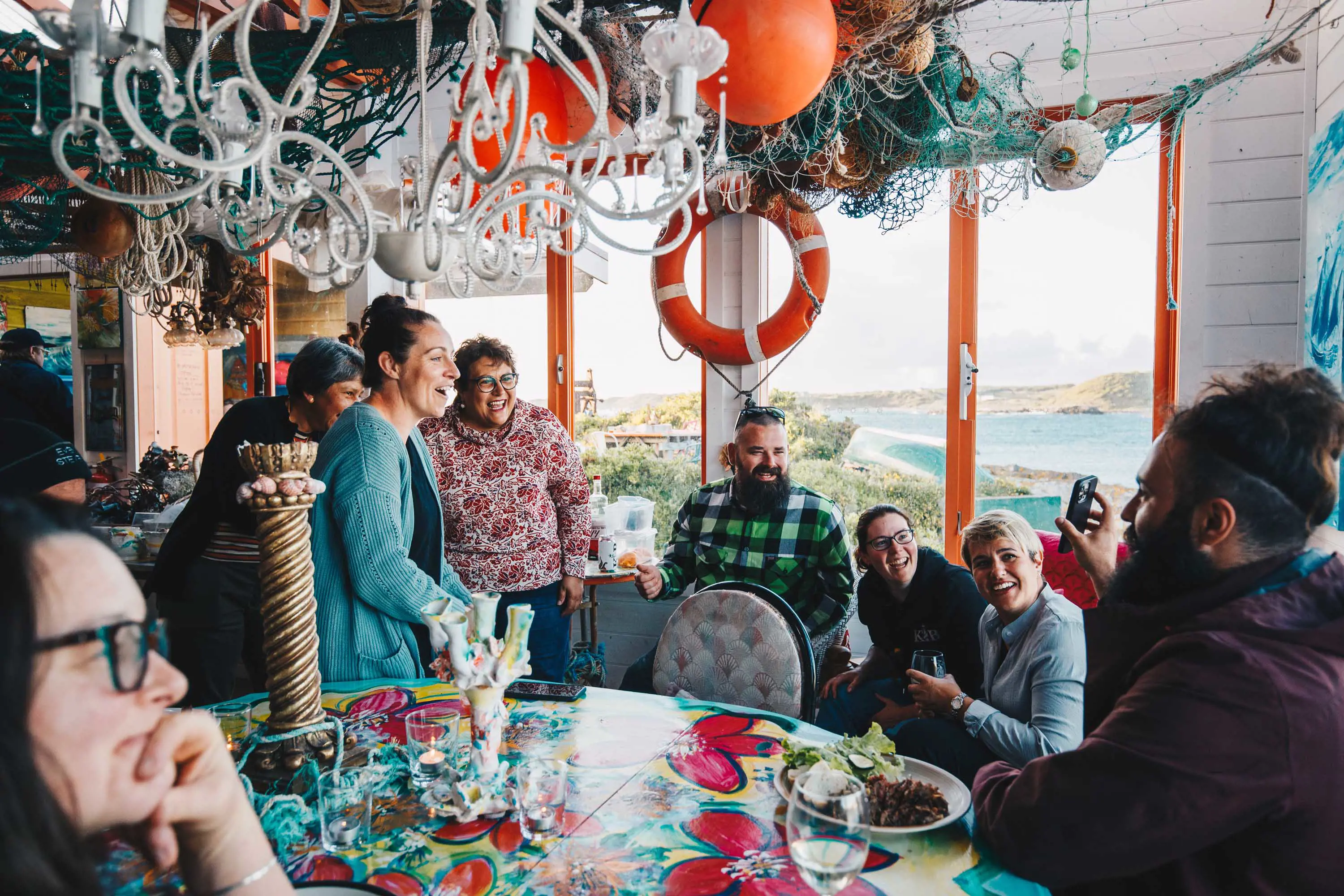 A large group of people gather around a table inside a weatherboard building overlooking a bay.