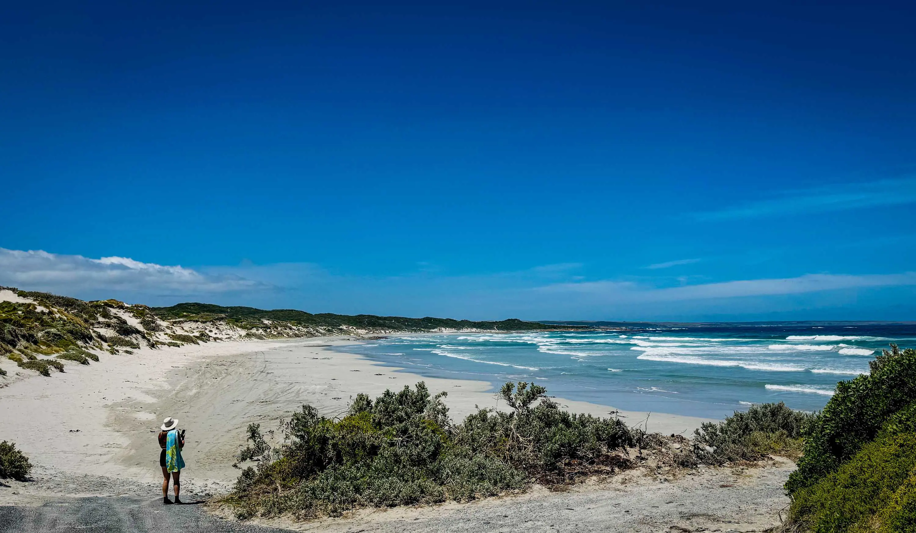 A person wearing a wide-brimmed sun hat stands at the edge of a beach and looks down the white sands to the water.