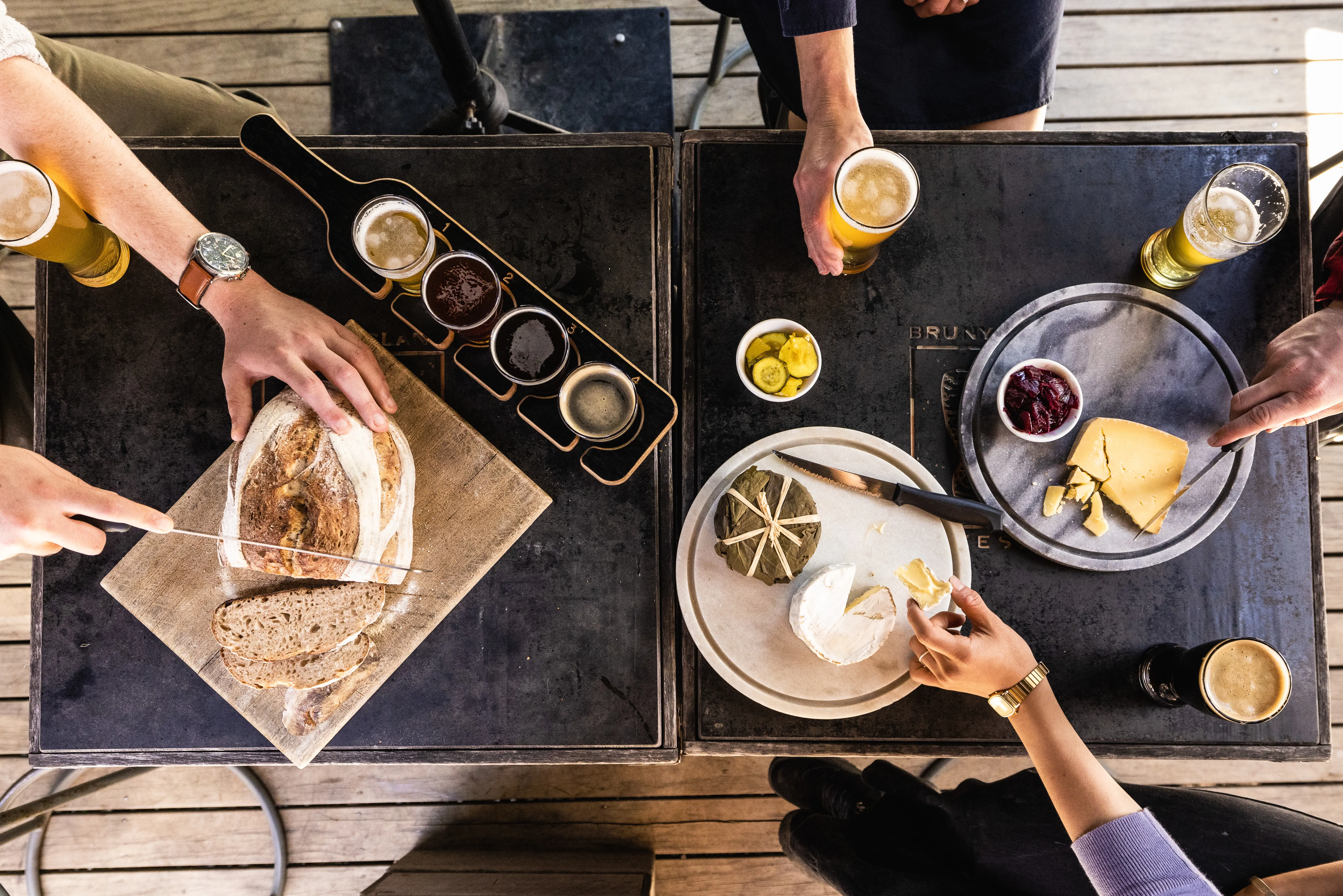 A group of friends sit down at a couple of small, square tables and enjoy beer and cheese with a large loaf of stone baked bread.