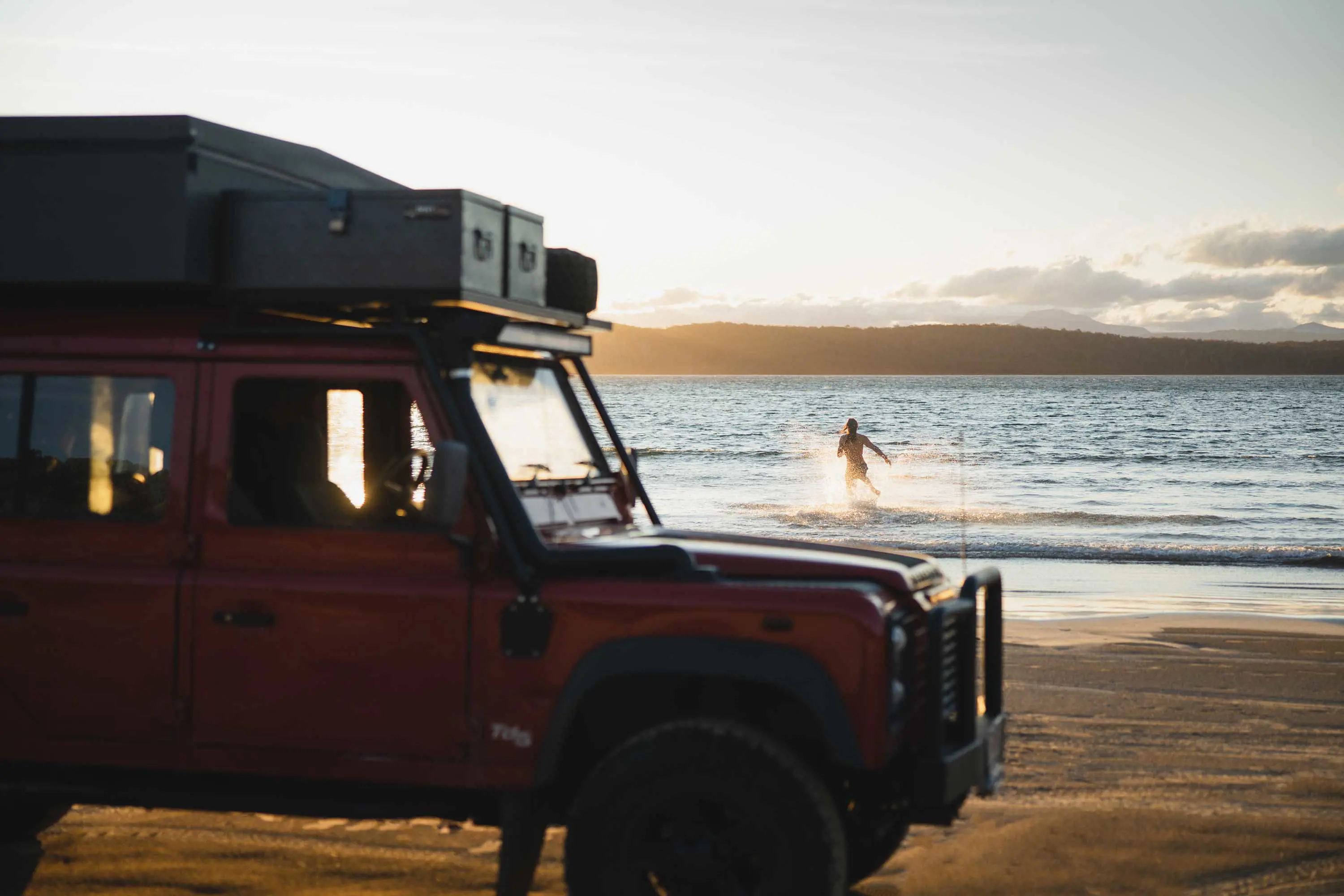 A large 4wd stacked with equipment is parked on sand in the foreground. A person splashes in knee high water in the background.