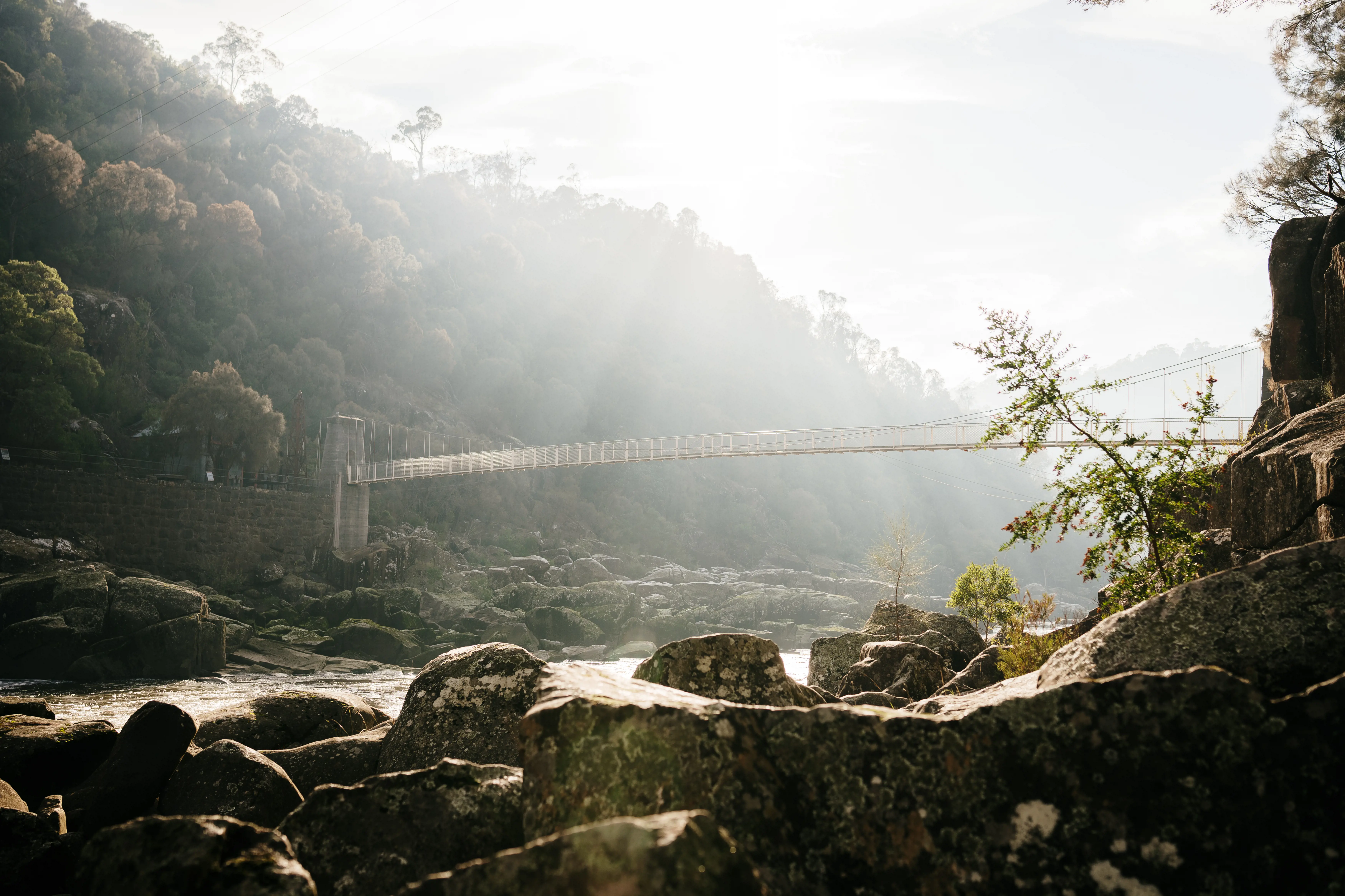 A long, thin bridge extends over a rocky river. Sunlight streams through clouds overhead.