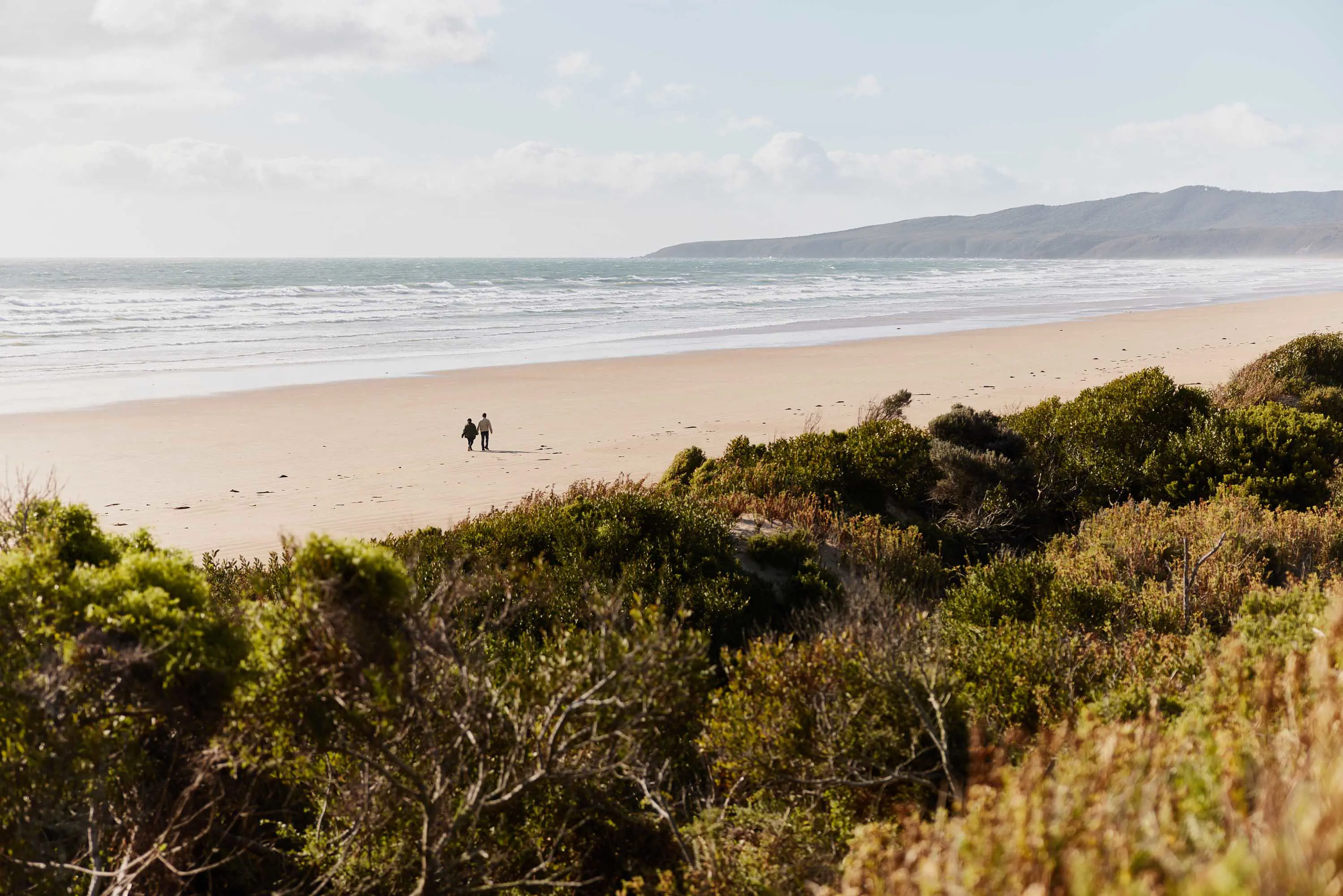 A couple walk along a wide beach of light yellow sand with small waves crashing into the shore. 