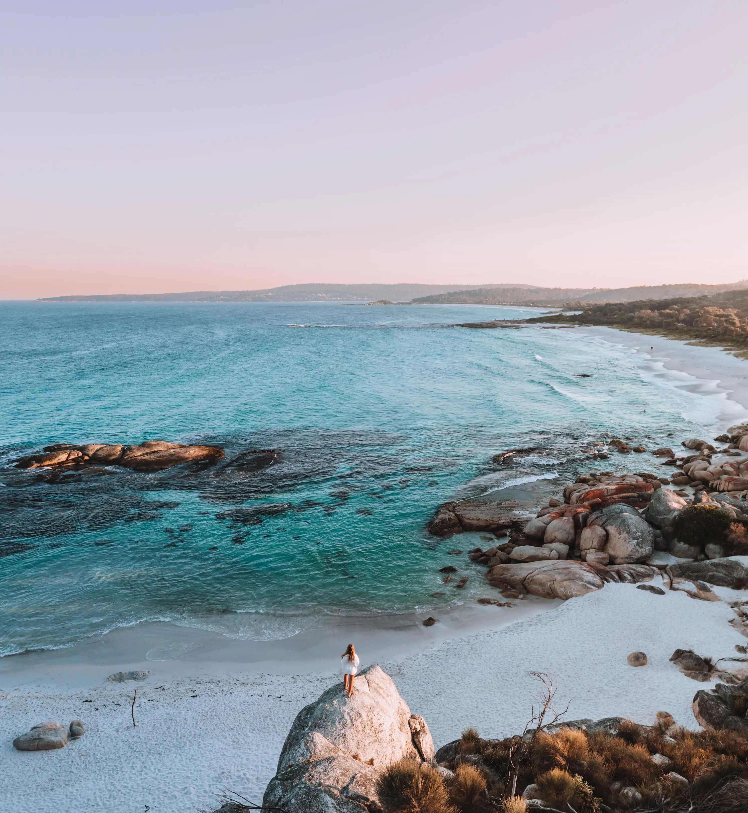 A woman wearing white clothing stands on top of a large rock overlooking white sandy beach and turquoise waters. 