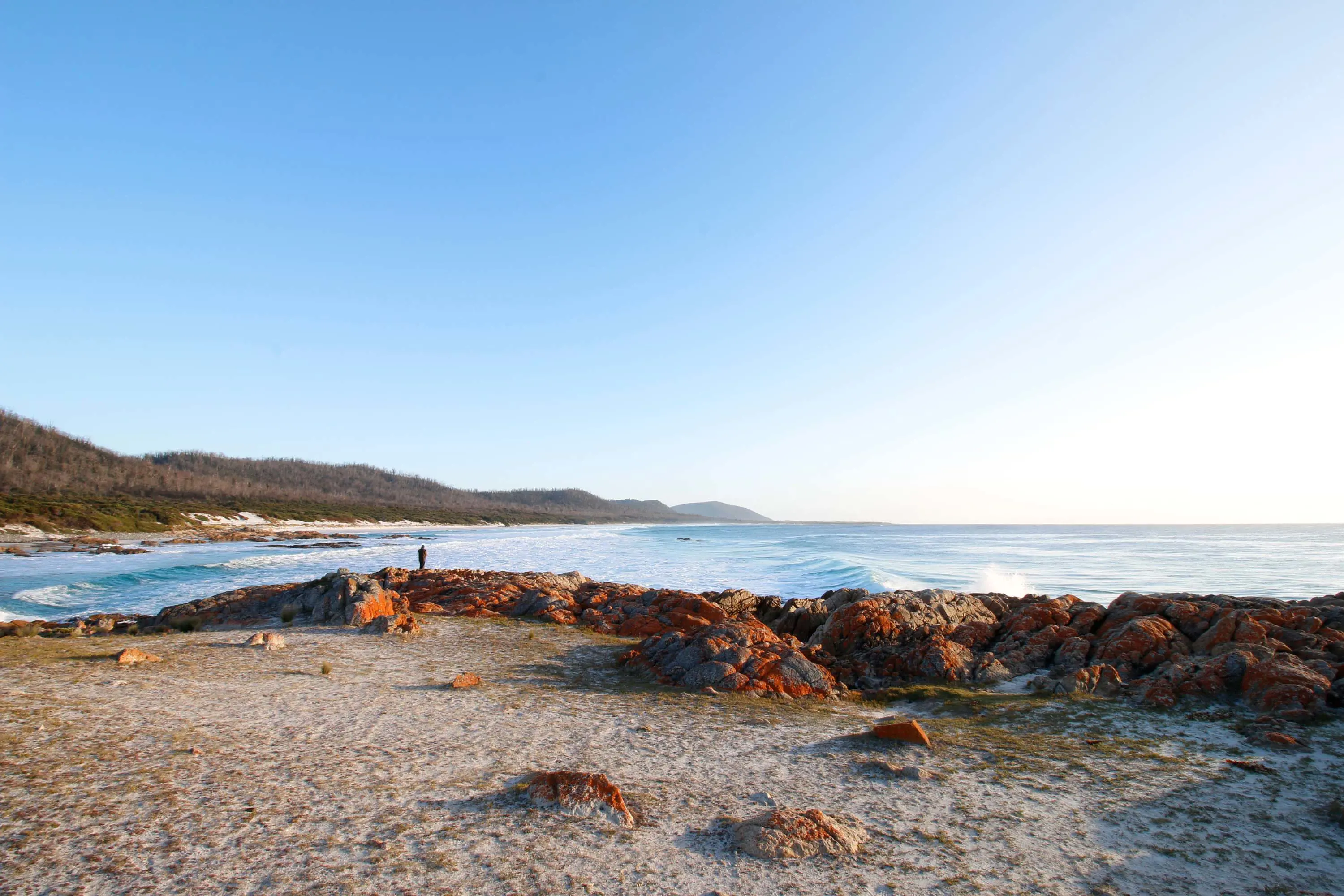 Rocks covered in orange lichen surround a clear blue beach with rolling waves, and a sun flare highlighting the sunset.