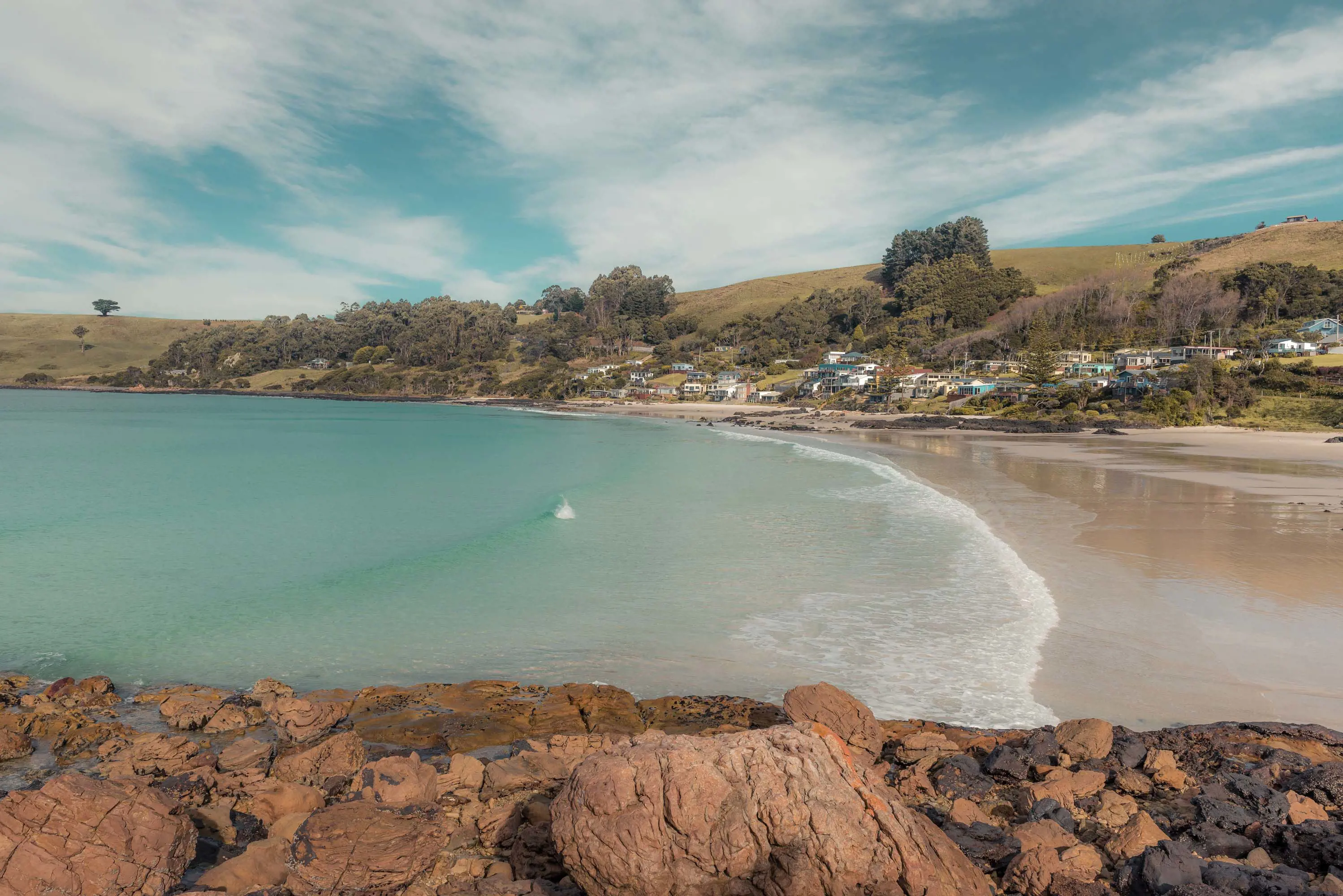 A sheltered beach, light aqua blue water lapping at the shore. On one side is a stretch of land with buildings on it, on the other some warm red and orange rocks.