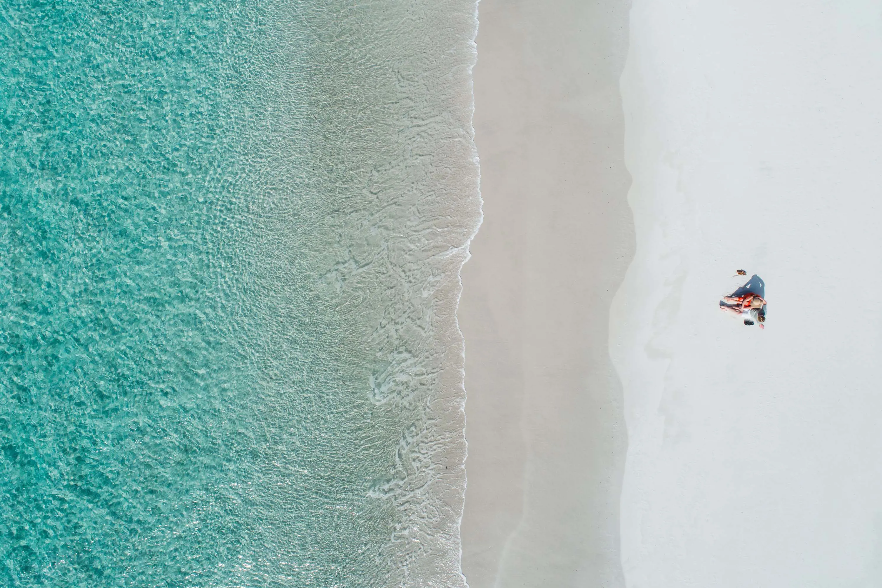 A striking aerial photo of the waterline at a beach. The water is crystal clear aqua blue, and two small figures relax on the white sand.