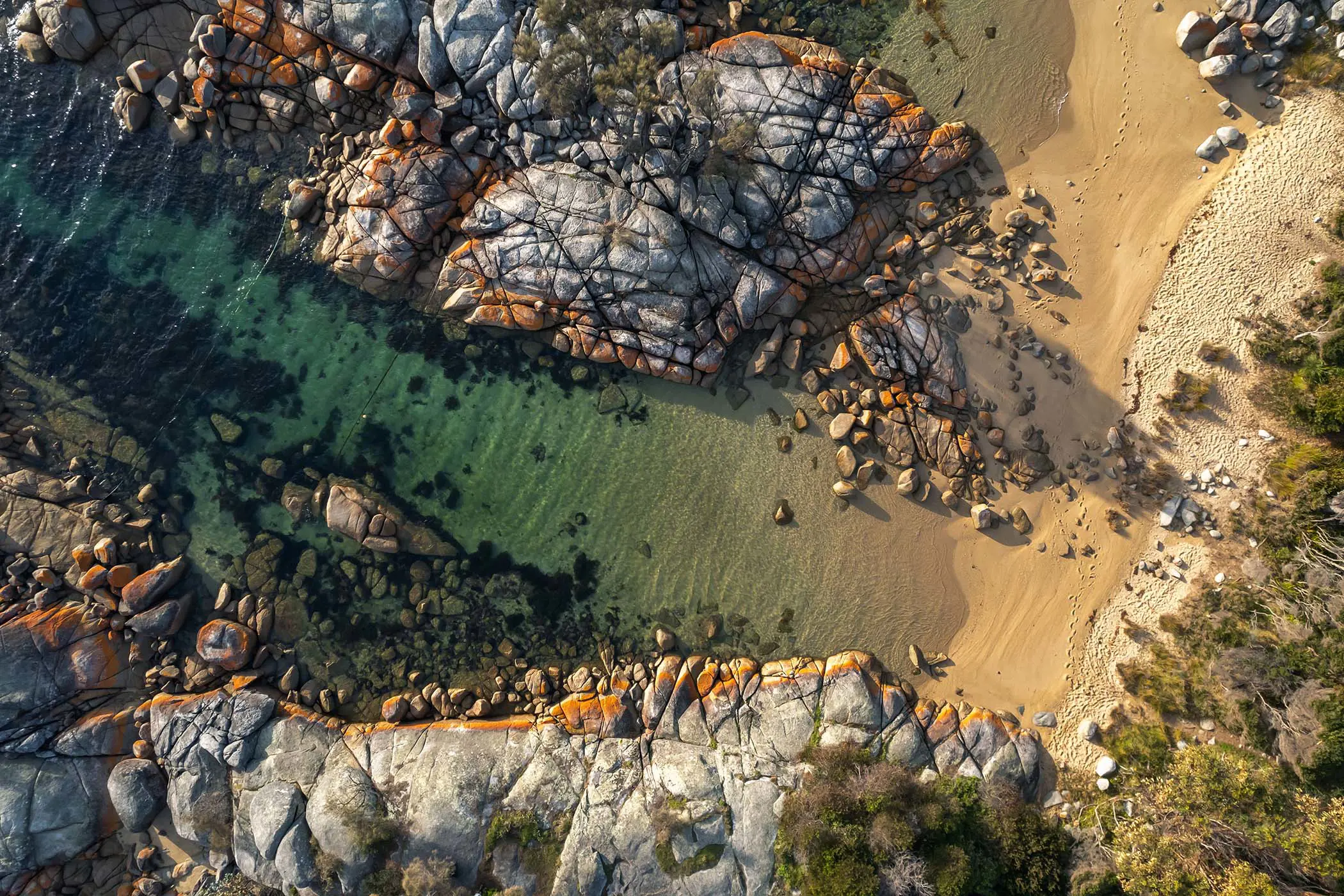 A birds-eye view of a striking, rock-strewn beach. The water is clear and bright and the grey granite rocks are splotched with orange streaks.