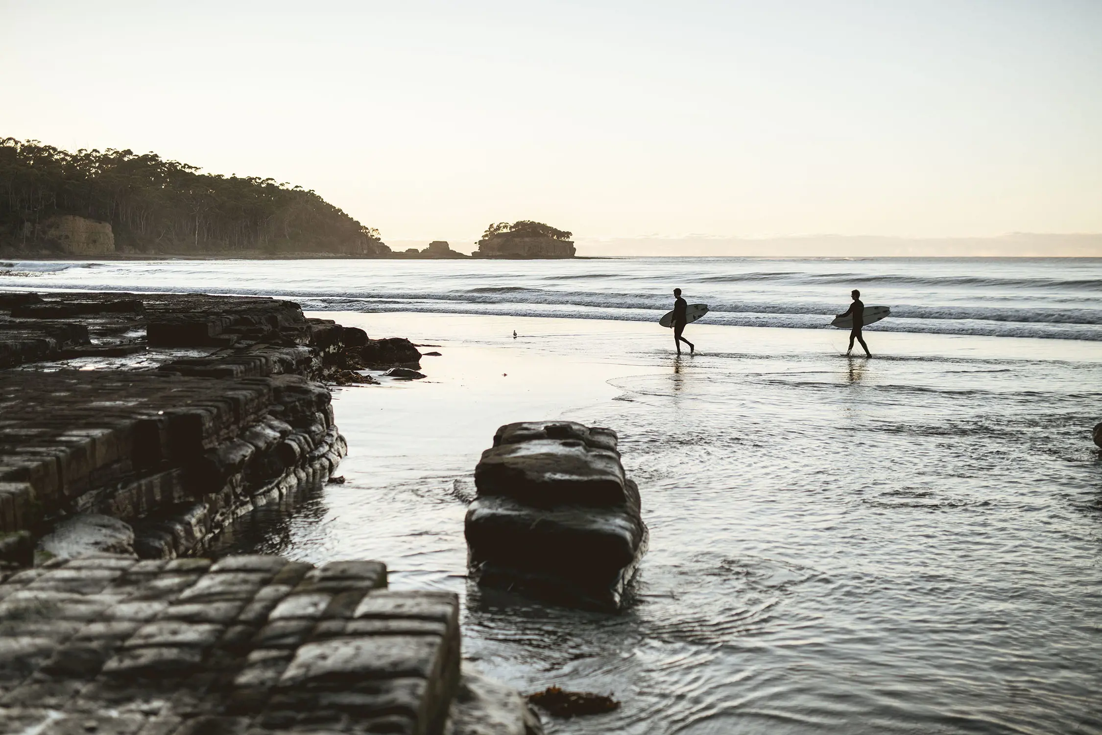 In the low light of sunset, two silhouettes carrying surfboard run through ankle-deep water. To the side, the ground is covered with rocks in a strange block-like formation.