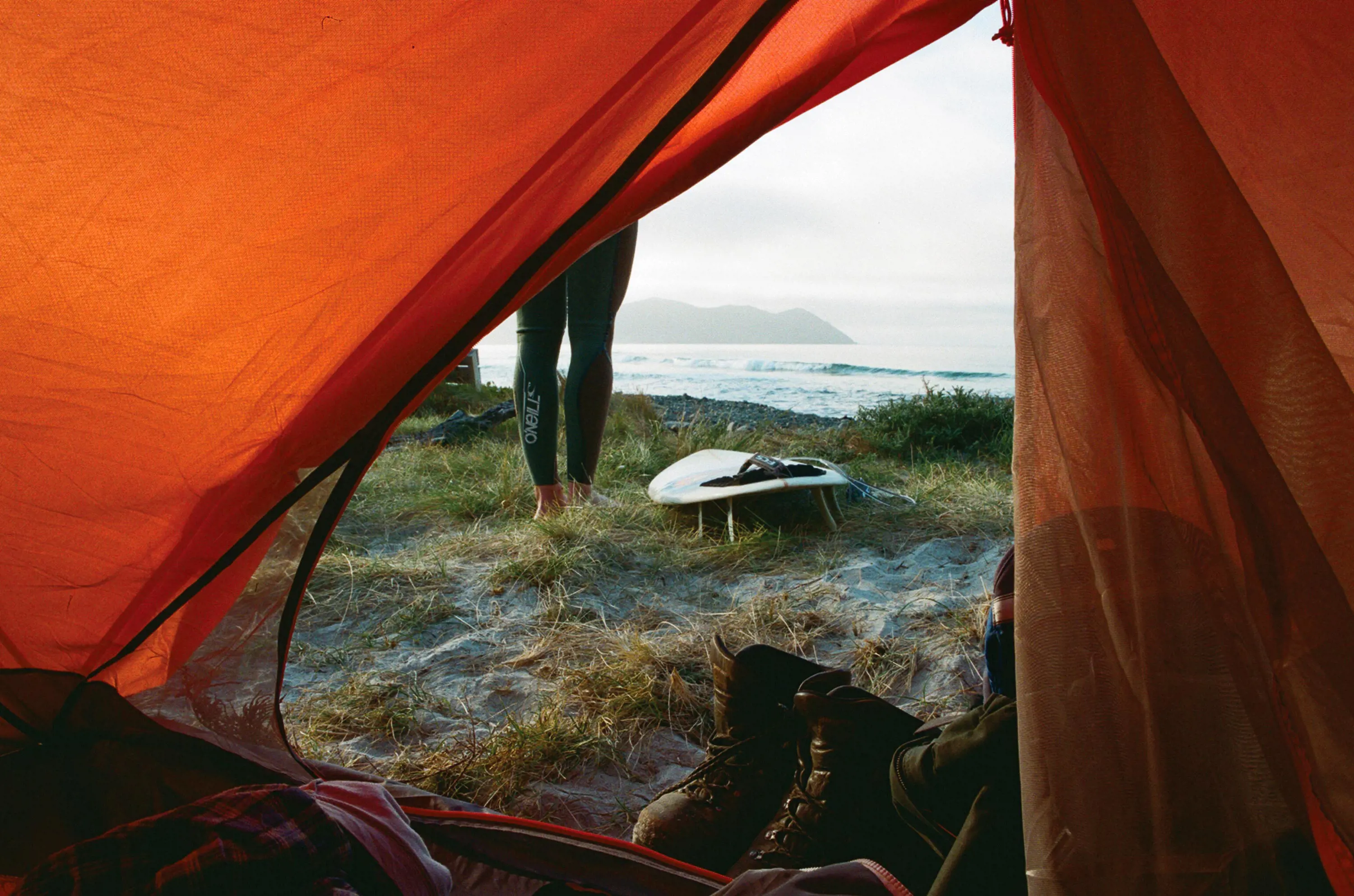 A man wearing a wetsui stands next to a surfboard lying on short grass and sand, seen through the opening of a orange tent.