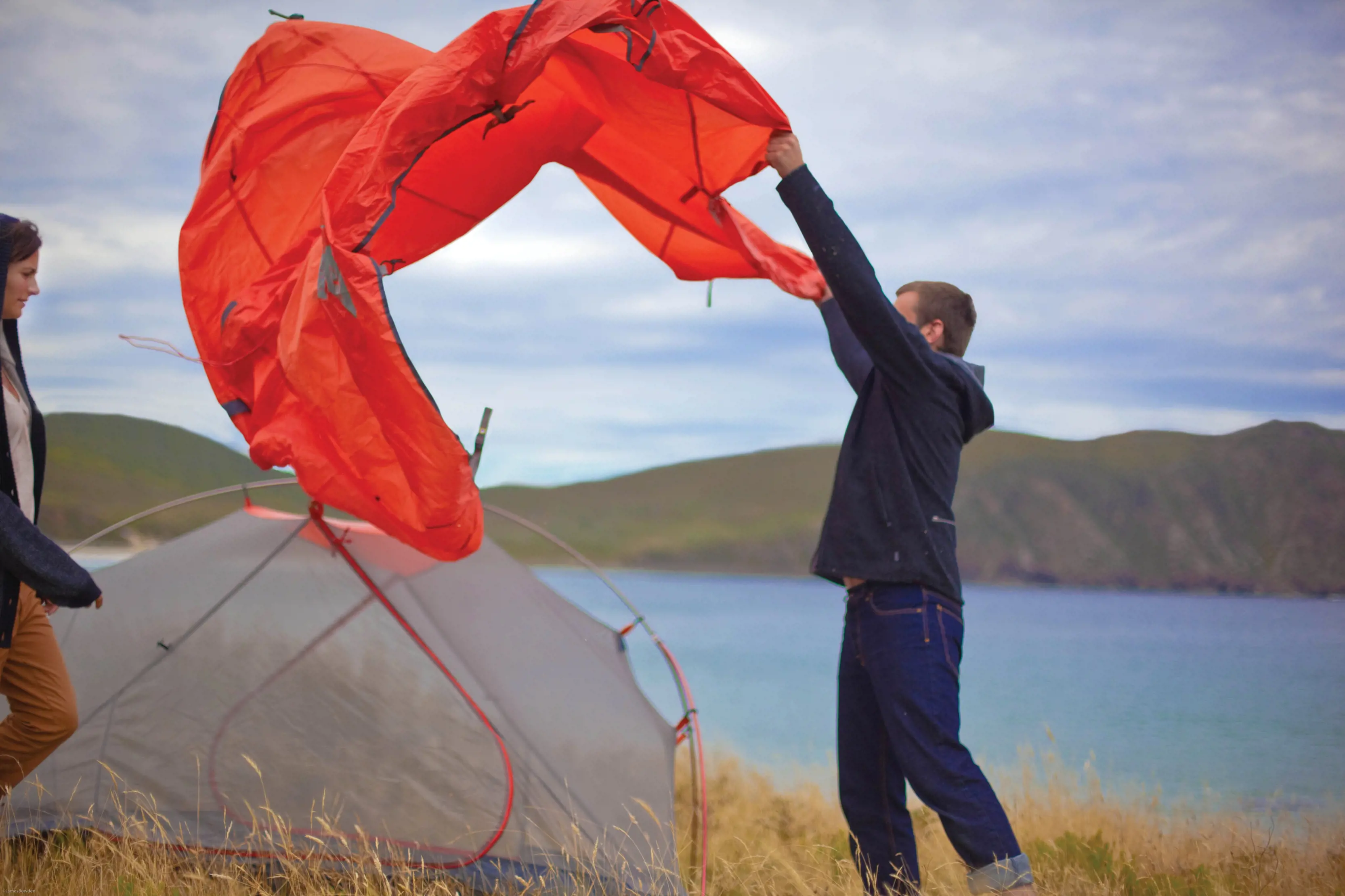 Exterior of people setting up camp near the Cape Bruny Lighthouse, there is water and mountains in the backdrop.