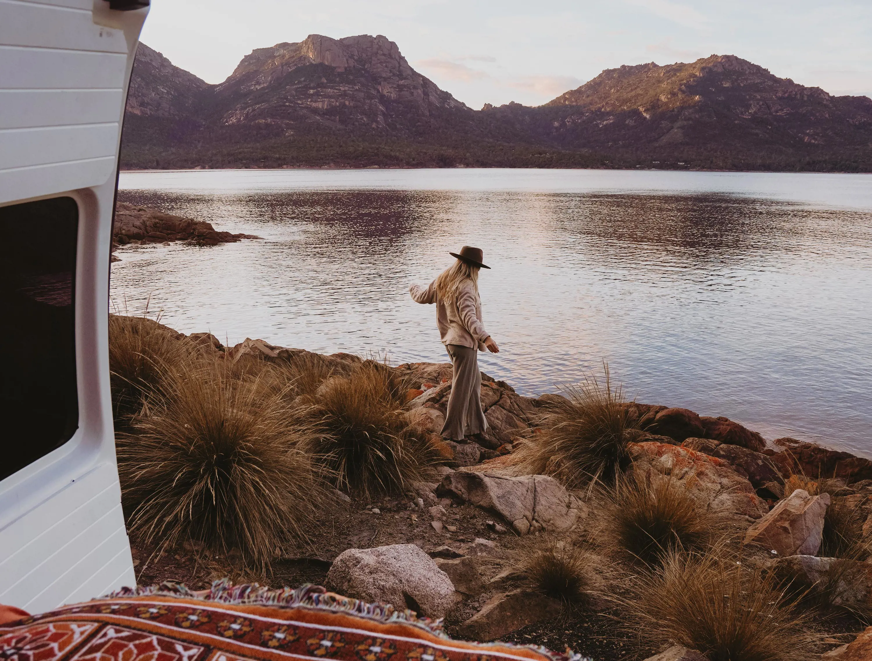 A woman wearing a wide-brimmed ha and warm clothing walks along rocks towards water with a mountain range in the distance.