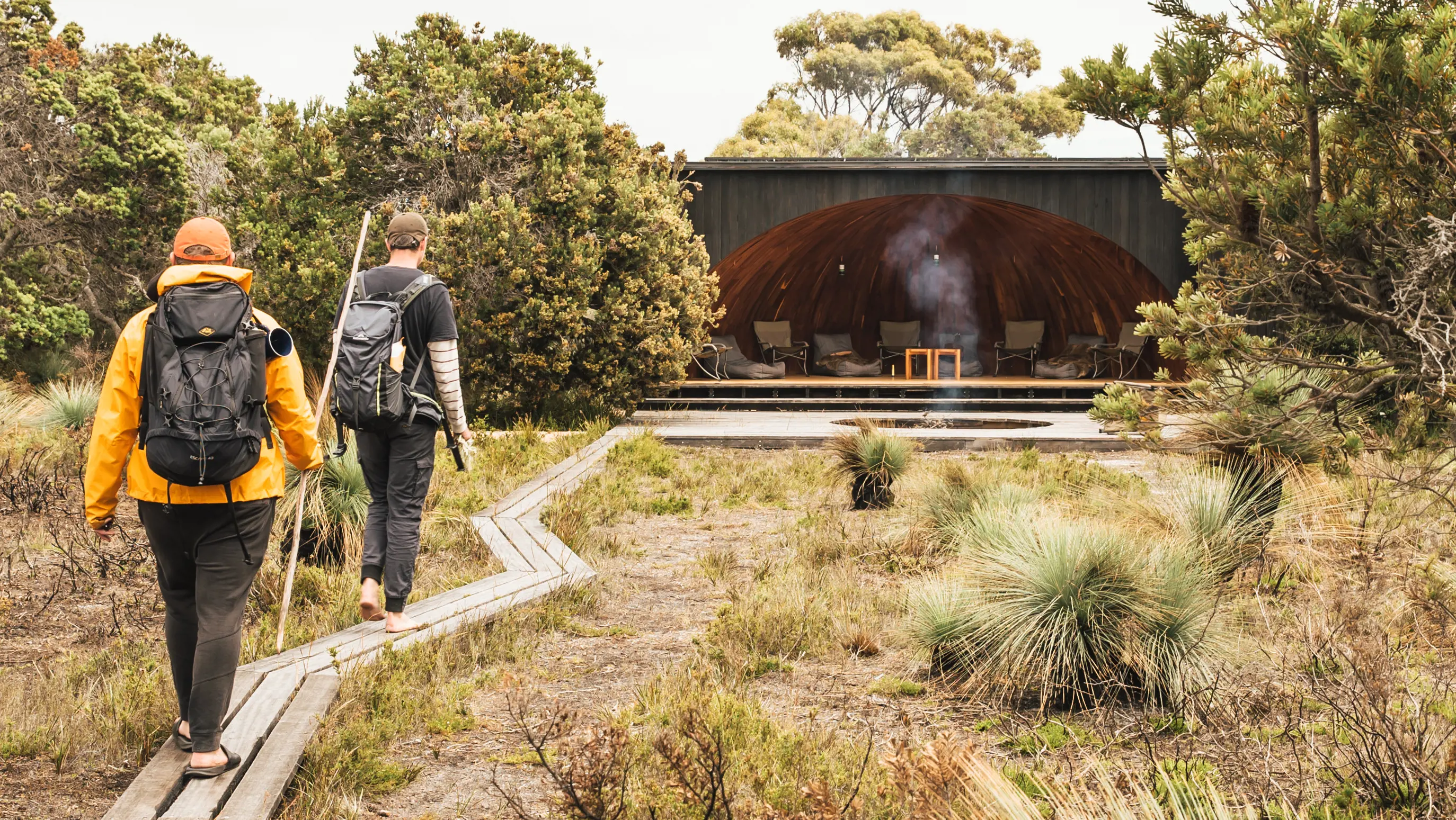 Two hikers walk along a wooden boardwalk, amongst the bush towards a campsite. Part of the Wukalina Walk.