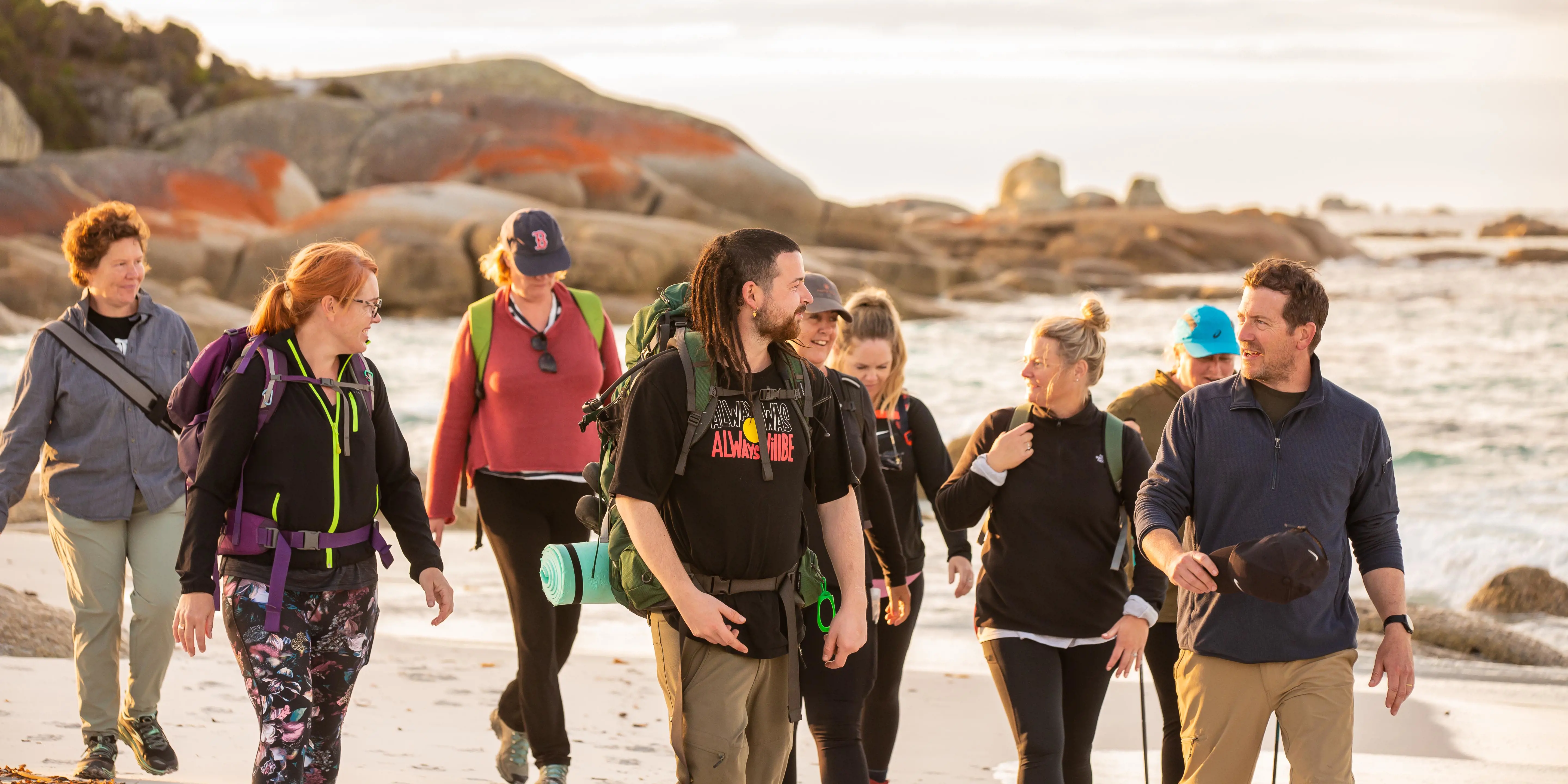 A group of people are led by a guide across white sands in front of orange rocks on a beach.